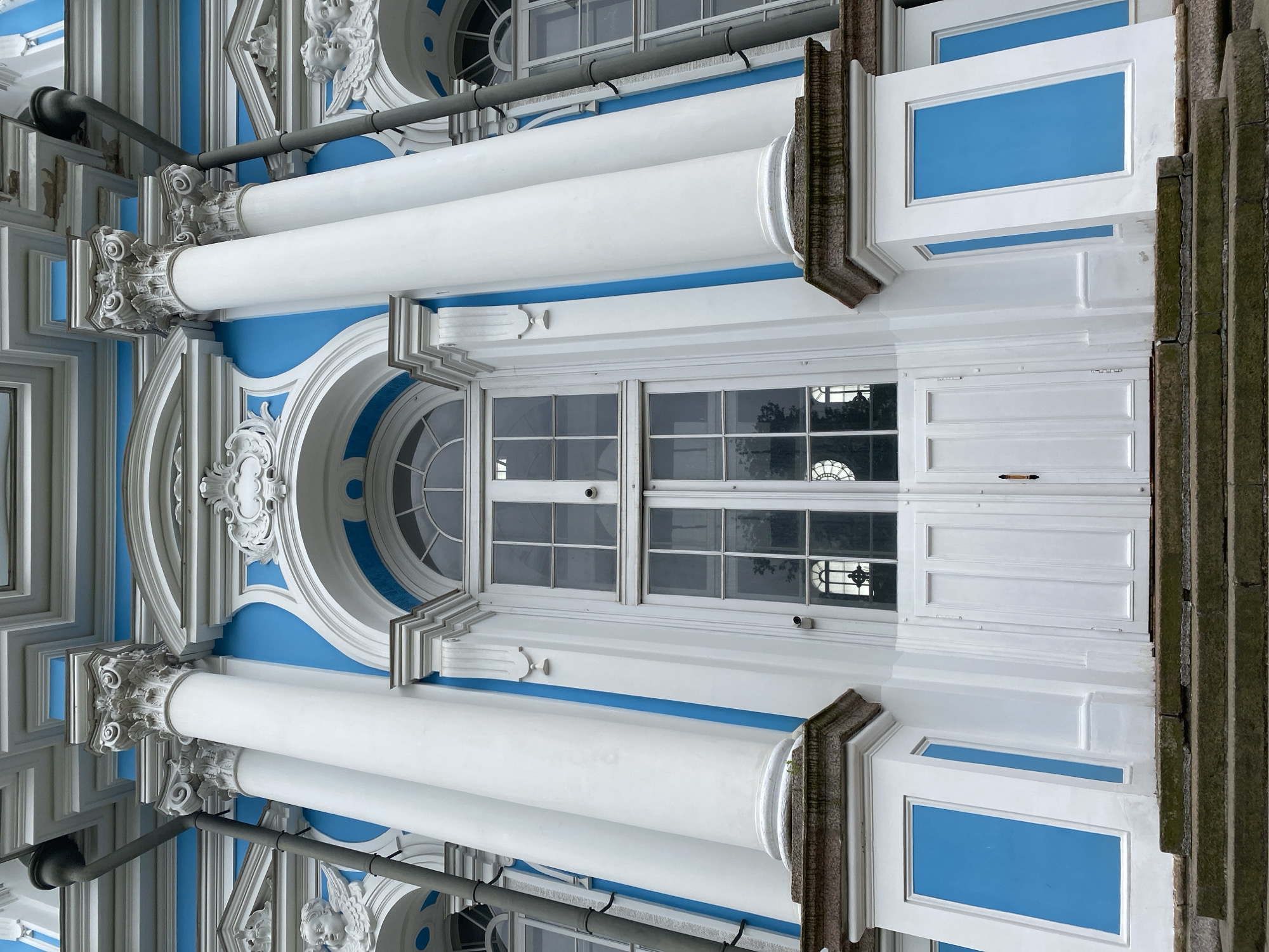 The image shows a close-up, low-angle view of the facade of the Smolny Cathedral in Saint Petersburg, Russia. The cathedral is painted in bright blue and white, with ornate architectural details. Dominating the center of the frame is a large, white double door, topped by a Palladian window with arched panes. Flanking the door are massive white columns with Corinthian capitals, set against the vibrant blue walls of the building. Decorative cherub sculptures are visible on either side of the central section. The foreground consists of weathered stone steps leading up to the entrance, partially covered in moss. The sky is overcast, casting a soft light on the scene.
