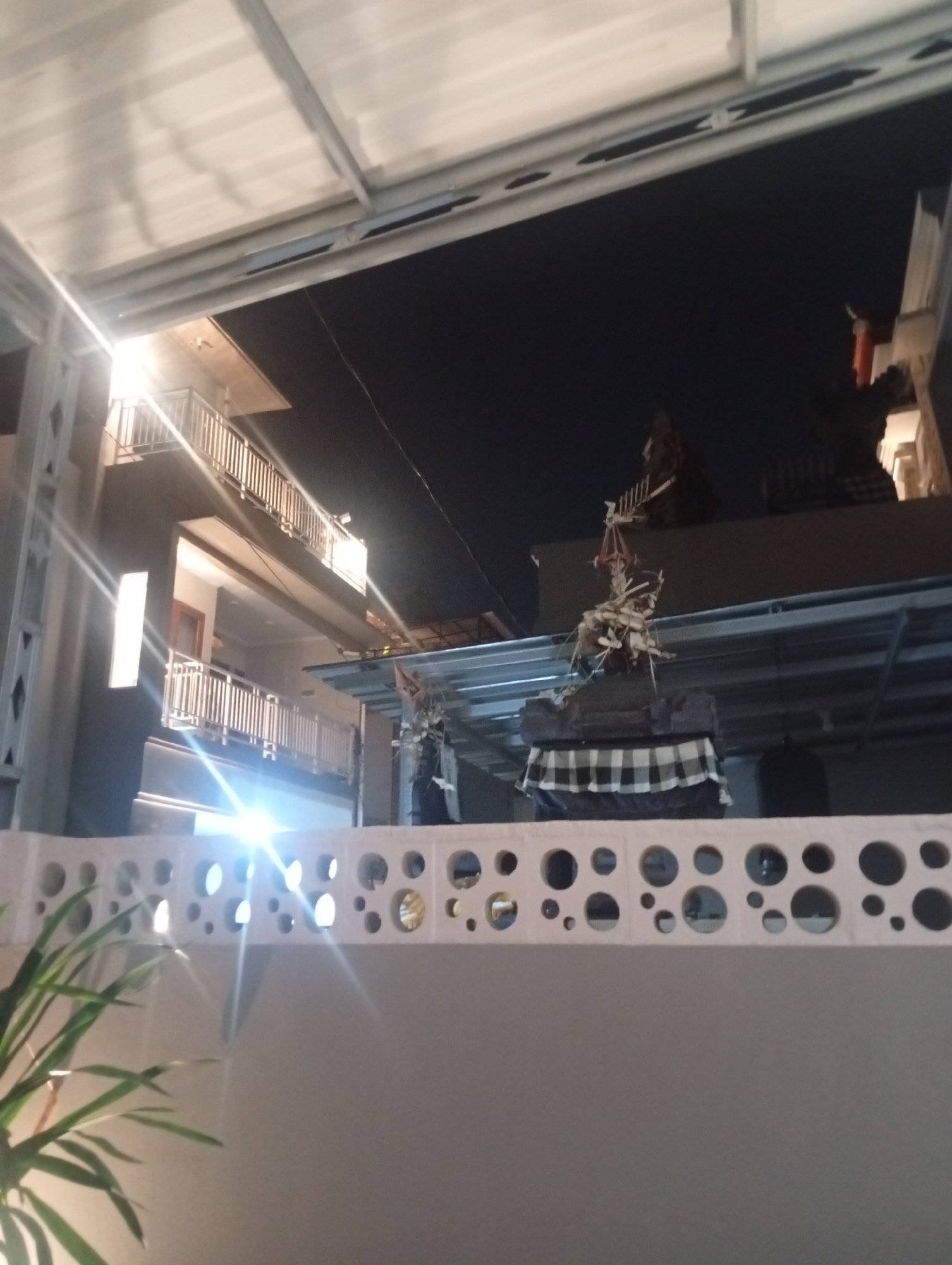 The image depicts an outdoor scene at night, likely in Denpasar, Indonesia, given the architectural style and the presence of what appears to be a religious offering or effigy. The foreground features a light-colored, decorative wall with circular cutouts, partially obscured by the fronds of a lush green plant on the left. Beyond this wall, a multi-story building with balconies and railings is visible, illuminated by bright lights that create lens flares. The building's architecture suggests a modern design, possibly a hotel or apartment complex. In the middle ground, a more elaborate structure stands, topped with what seems to be a traditional offering, wrapped in black and white checkered fabric. This effigy is made of what looks like wood, bamboo, and possibly other organic materials, giving it a rustic and spiritual appearance. The background is dominated by the dark night sky, with hints of other buildings or structures. The overall impression is one of a cultural or ceremonial event taking place in an urban environment.