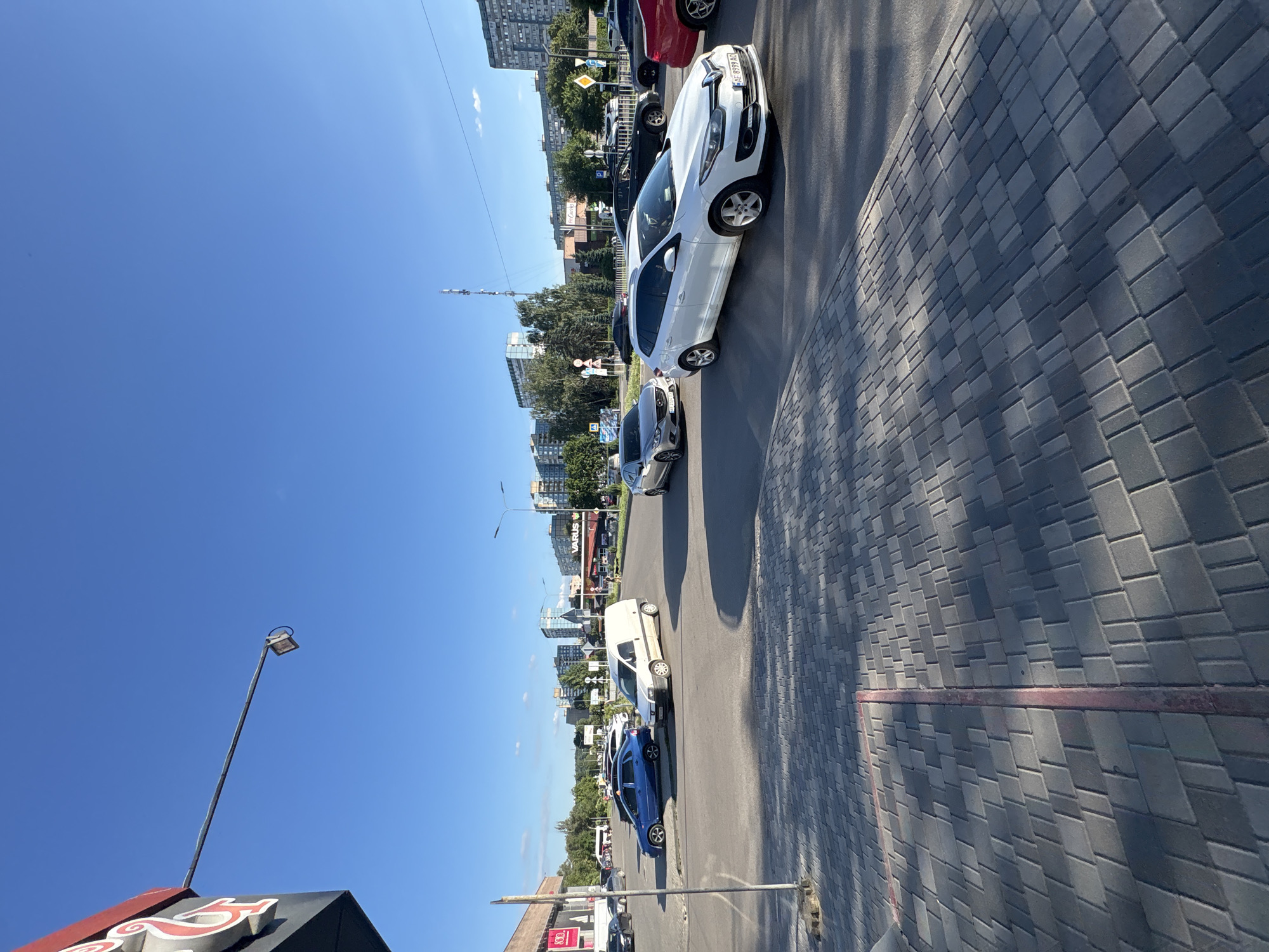 In Dnipro, Ukraine, on a clear, sunny day, a wide street intersection is depicted with multiple vehicles. The foreground shows a paved pedestrian area with interlocking gray pavers, marked by a red line. Across the street, several cars are visible, including a white Renault station wagon with license plate AE 8999 AQ, and a white van. A blue sedan is parked on the left side of the street. In the background, various buildings, including apartment complexes and commercial structures with signage such as "VARUS," line the horizon. A radio tower stands tall in the distance. The overall scene is characteristic of a busy urban environment.