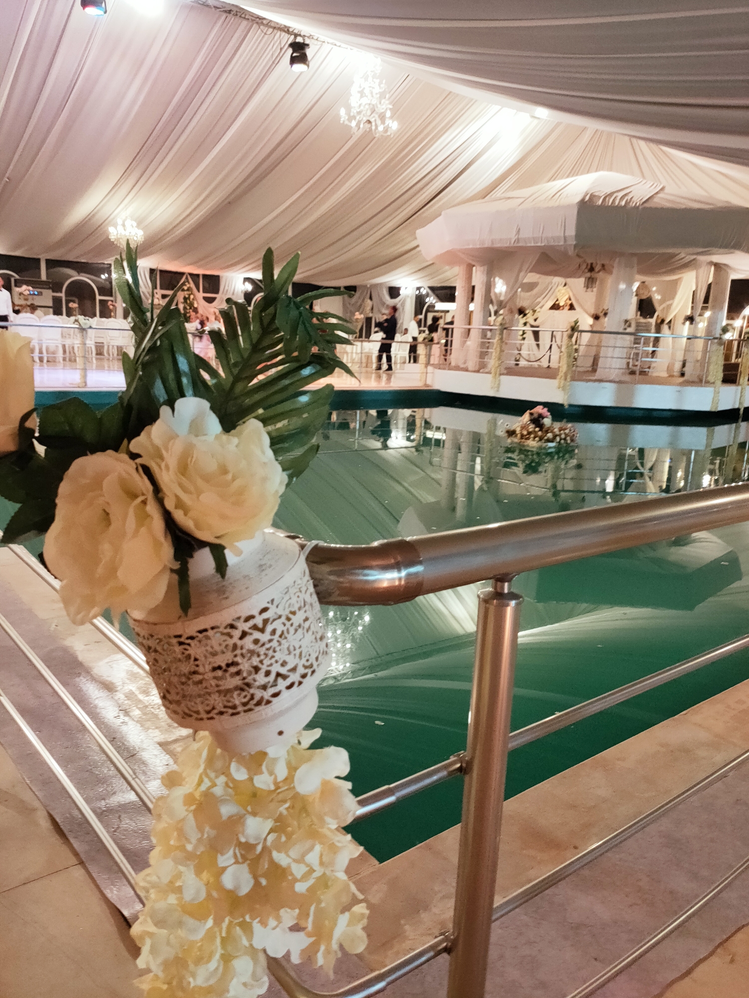This image depicts a lavish indoor event space, likely a banquet hall or reception venue, located in Mornag, Tunisia. The space is covered by a white draped tent ceiling with ornate chandeliers. A prominent feature is a large, turquoise-colored pool, which appears to be the central focus of the venue. A white gazebo-like structure with draped fabric stands over the pool.
In the foreground, a metal railing separates the viewer from the pool. Decorative elements adorn the railing, including a white, intricately carved lantern holding pale yellow roses and greenery, and a cascade of pale yellow artificial flowers hanging below. The floor in the immediate foreground appears to be tiled.
In the background, tables and chairs are set up, suggesting a dining or seating area. Several people are visible, some standing near the tables and others interacting in the background. The overall ambiance is one of elegance and festivity, suggesting a special occasion such as a wedding or a formal celebration.