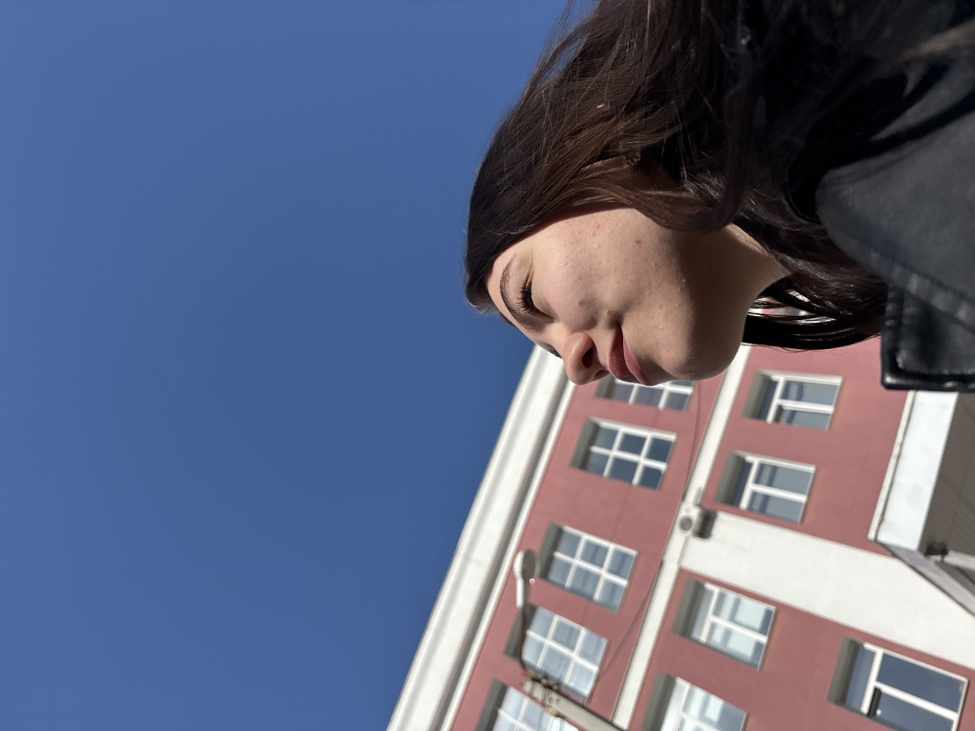 A low-angle photograph shows a woman's face against a backdrop of a building and blue sky in Perm, Russia. The woman has dark hair and fair skin, with visible blemishes on her face. She is wearing a dark-colored jacket. The building is painted in red-brown, with white window frames and white trim. The windows are rectangular with white grid patterns. The sky is a clear, solid blue.