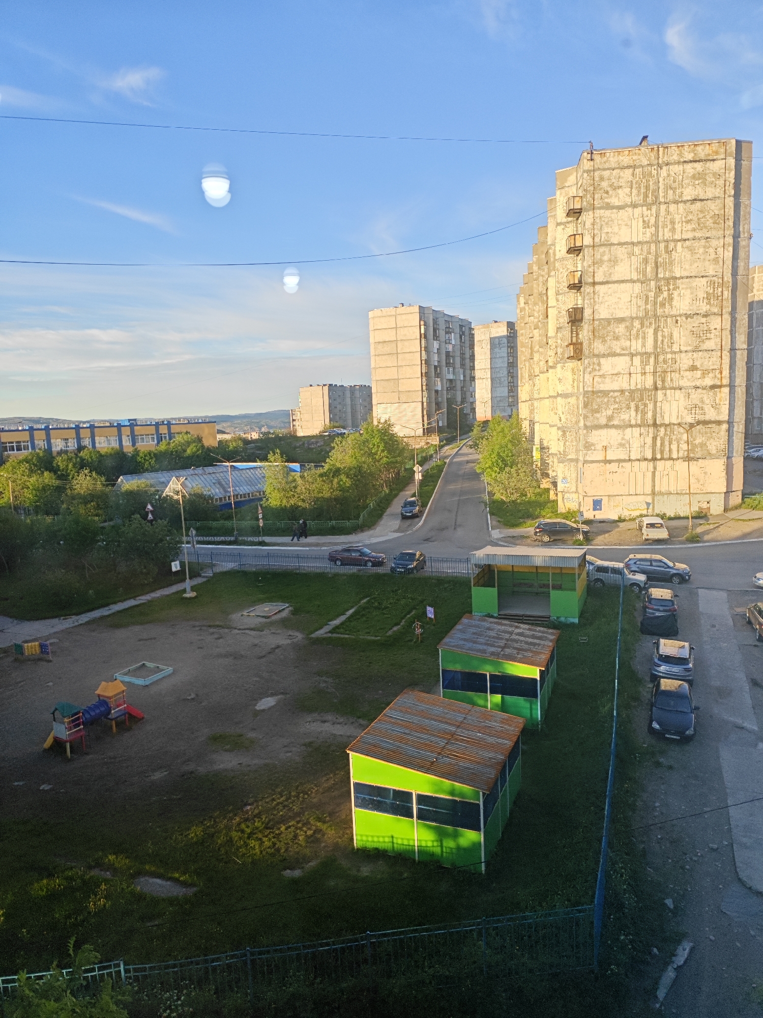 The image captures a sun-drenched urban scene in Polyarny, Russia, dominated by towering, weathered apartment buildings that cast long shadows. In the foreground, a children's playground with a sandbox and a colorful climbing structure sits on a patch of uneven dirt and grass. Adjacent to the playground are two identical, open-sided green shelters with corrugated metal roofs, partially illuminated by the low-angled sunlight.
A paved road winds through the middle ground, with several parked cars lining its edge and a few vehicles in motion. Two figures, appearing to be pedestrians, walk along the sidewalk. Beyond the road, a row of lush green trees separates the residential area from a modern, light-colored building with blue trim, likely a school or public facility. In the background, more apartment blocks rise against a clear blue sky, with faint wisps of clouds. Two unusual, bright, spherical lens flares are visible in the upper sky, likely caused by the sun reflecting off the camera lens. The overall impression is one of a typical summer day in a Soviet-era housing development.