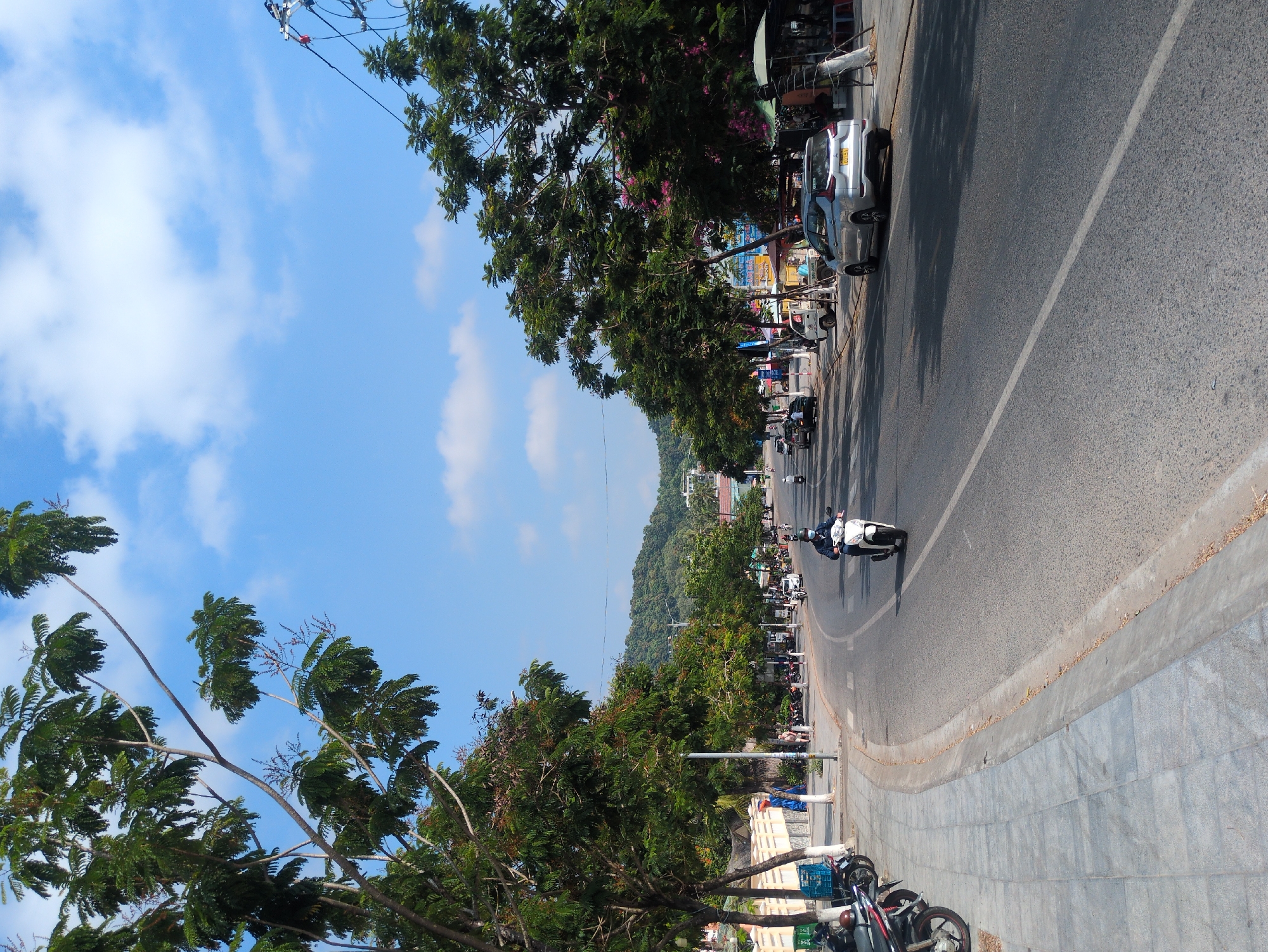 The image depicts a street scene in Phú Quốc, Vietnam, showcasing a road curving towards the horizon, bordered by sidewalks and trees. A white-helmeted person rides a scooter down the road, which also shows other vehicles. Cars and motorcycles are parked along the right side of the road. Trees line both sides of the road, and a hill is visible in the distance under a blue sky with some clouds. Power lines can be seen above the street on the right side.