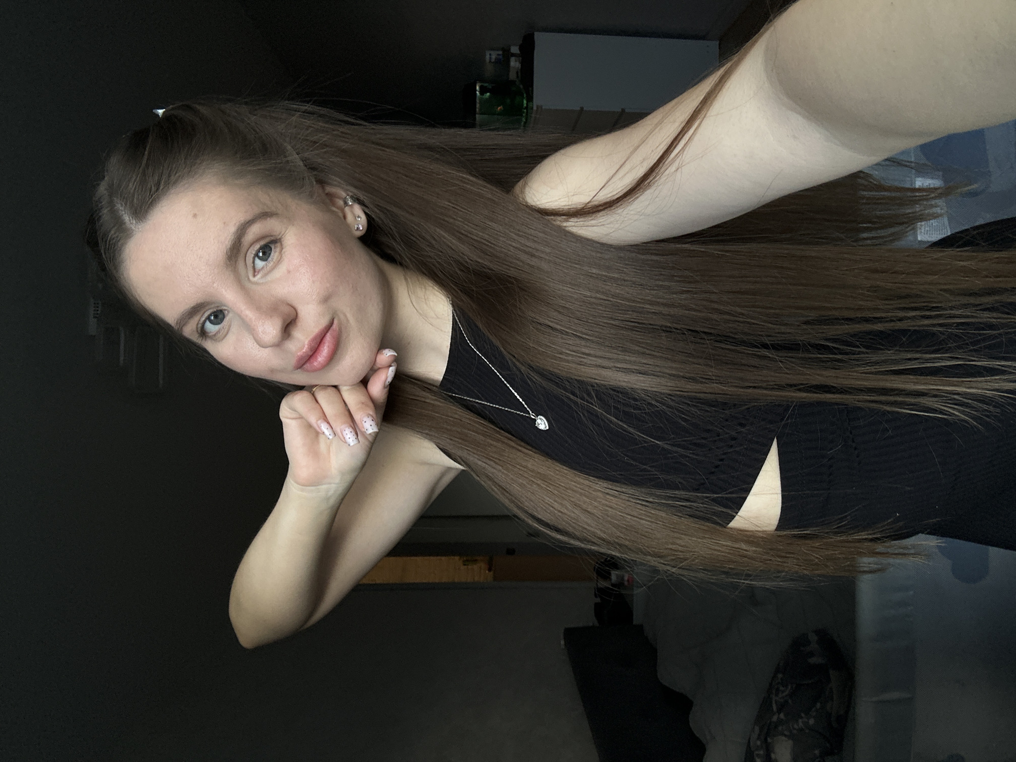 A young woman with long brown hair partially pulled back, wearing a black top and a silver necklace, poses for a selfie. Her nails are decorated with a white and black polka dot pattern. The background is a dimly lit interior, with some indistinct objects visible. It is impossible to ascertain whether the image was taken in Klin, Russia, based on the visual information available.