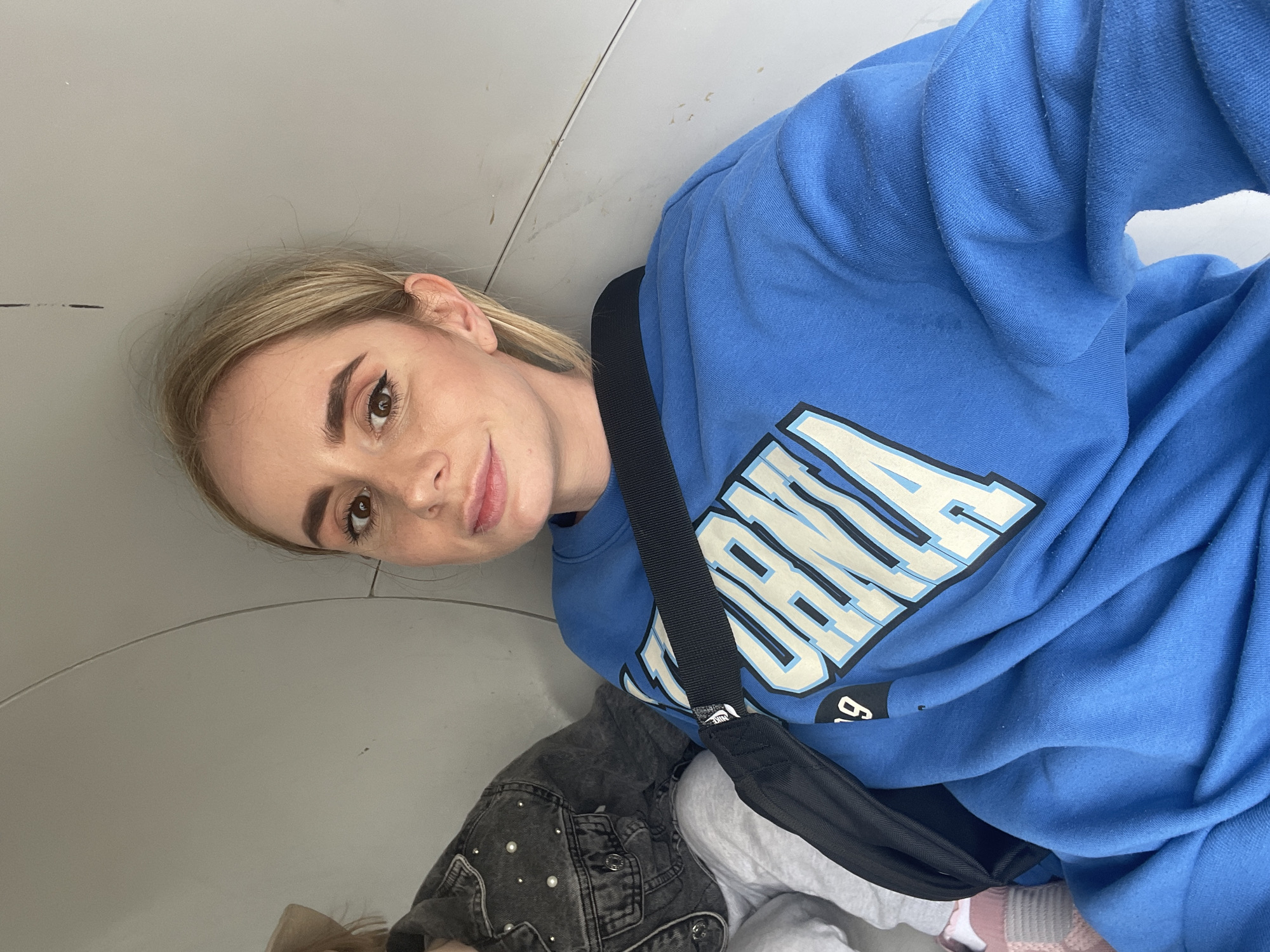 A blonde woman is posing indoors. She has fair skin, brown eyes, and arched eyebrows, and is wearing eyeliner. She is wearing a blue sweatshirt with white lettering and a black strap across her body. Parts of clothing in the lower section of the picture appear to be a gray denim jacket, white pants, and pink socks. The surroundings include a white wall. The location in Vladivostok, Russia is not discernible from the provided image.