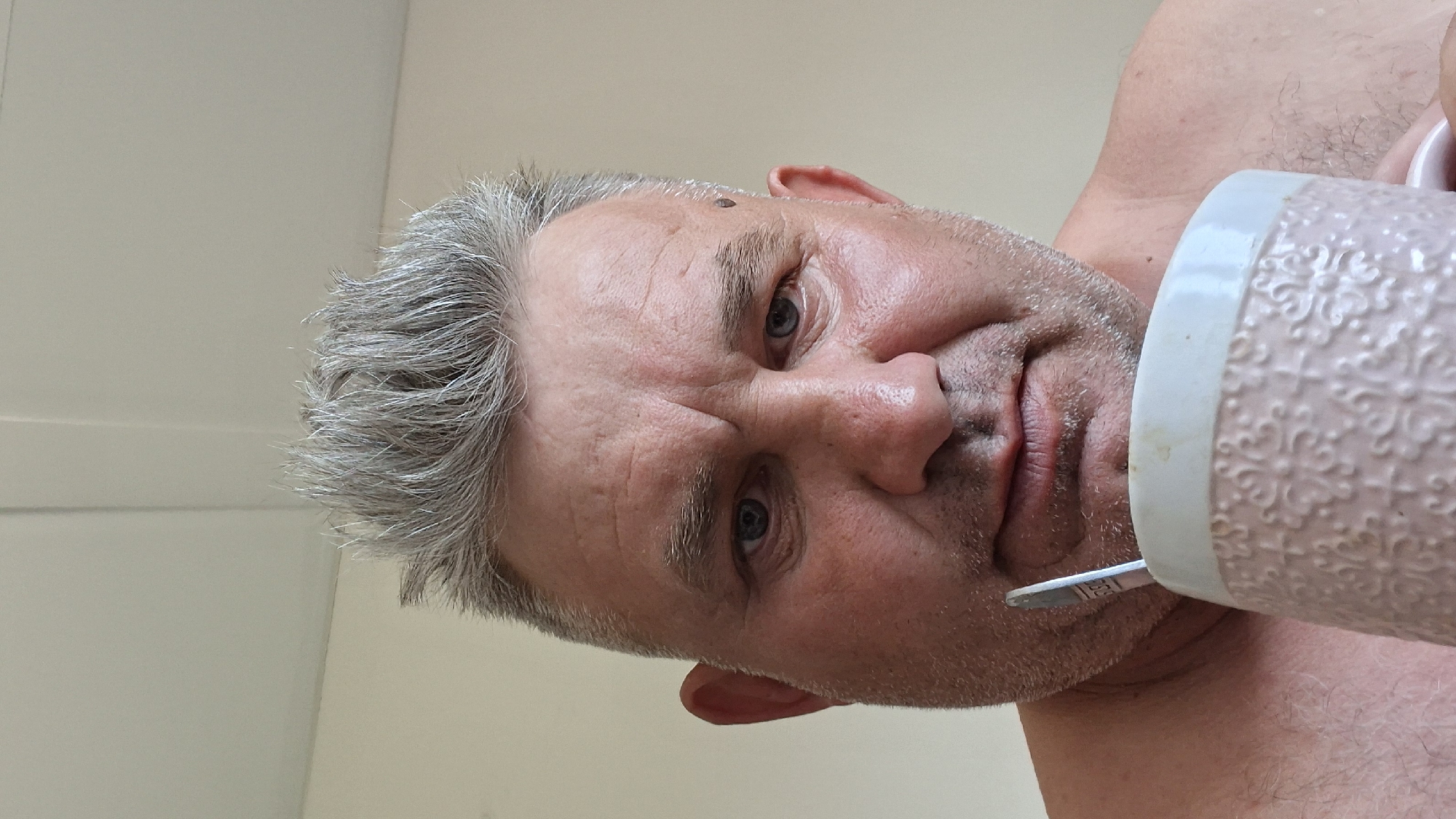 The media file is a close-up, low-angle, rotated photograph of a man holding a mug with a metal straw. The man has graying hair and is looking towards the camera with a neutral expression. He appears to be indoors, with a plain, light-colored wall visible in the background. The lighting suggests it is daytime. The man's upper body is partially visible, and he is wearing what appears to be a textured, light-colored mug. A silver metal straw is inserted into the mug. There is no visible text in the image. The scene is simple and focused on the man and the mug, with no other distinguishing features of the setting.