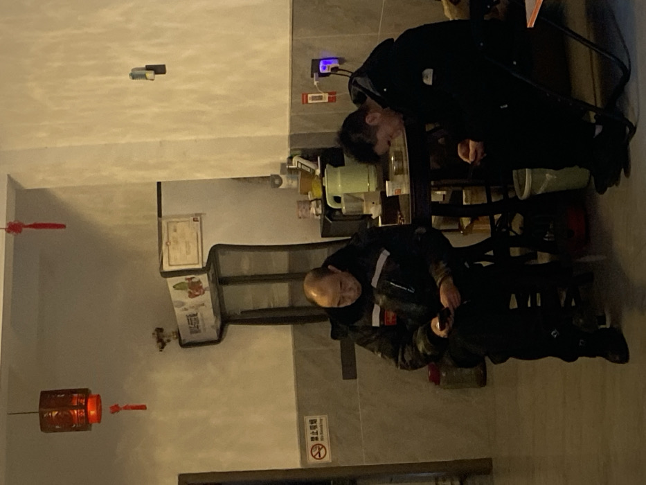 Two men in dark clothing are present in a dimly lit interior. One stands near a table with a green kettle and other items, while the other is seated with what appears to be a tool in his hand. Decorative lanterns hang on the left. Signs, possibly in Chinese, are visible on the wall. The room's walls are a light color.
