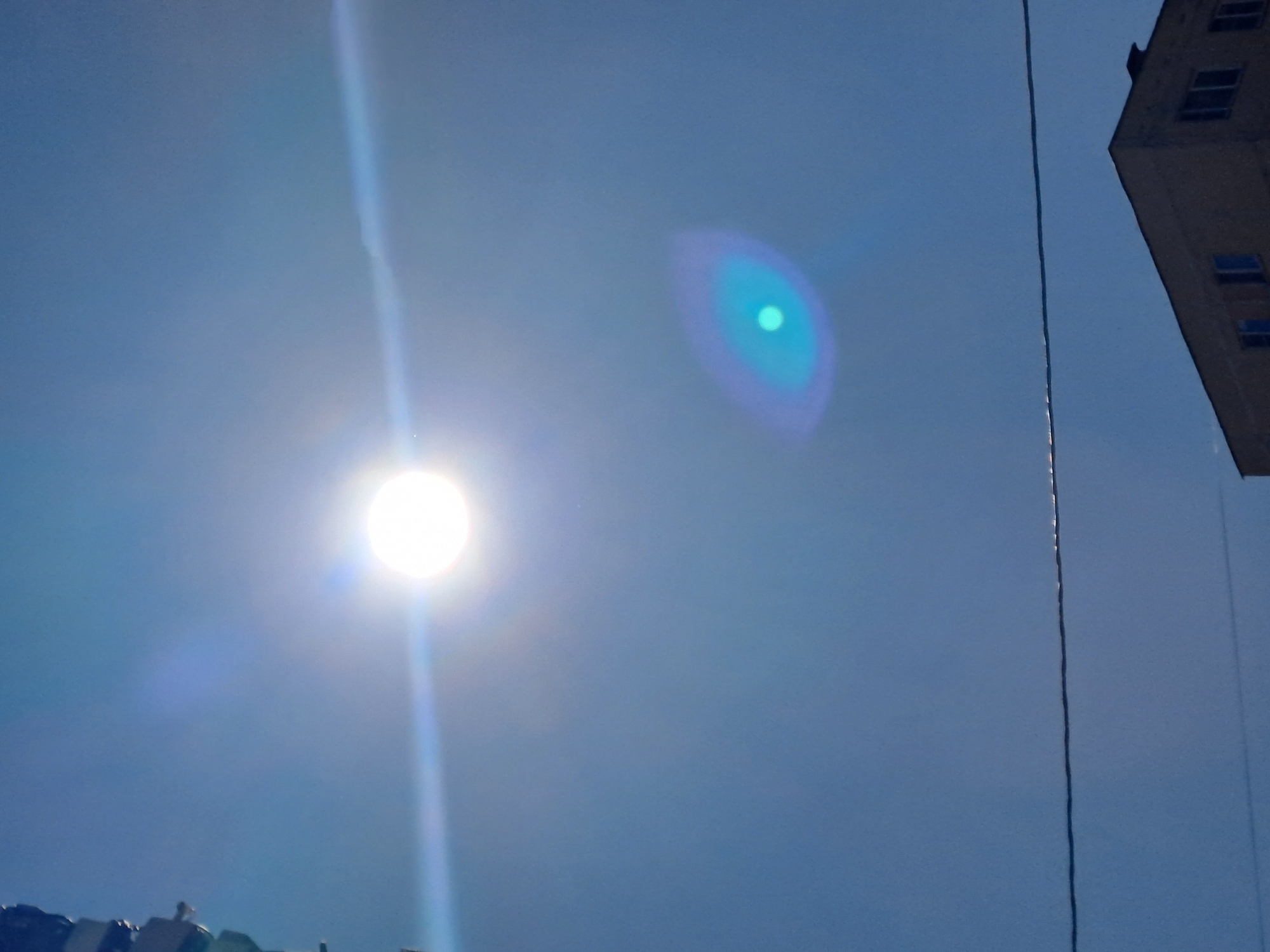 The image captures a bright sun low in a clear blue sky, with noticeable lens flare creating horizontal and oval light streaks. In the lower right quadrant, the corner of a multi-story building with multiple windows is visible. A thin, dark wire stretches horizontally across the mid-section of the frame, suggesting an urban or suburban setting. The upper left portion of the image shows a partial view of a vertical structure, possibly a utility pole or part of a building. The overall scene is devoid of any people or specific identifying landmarks that would pinpoint a location within Elektrostal, Russia.