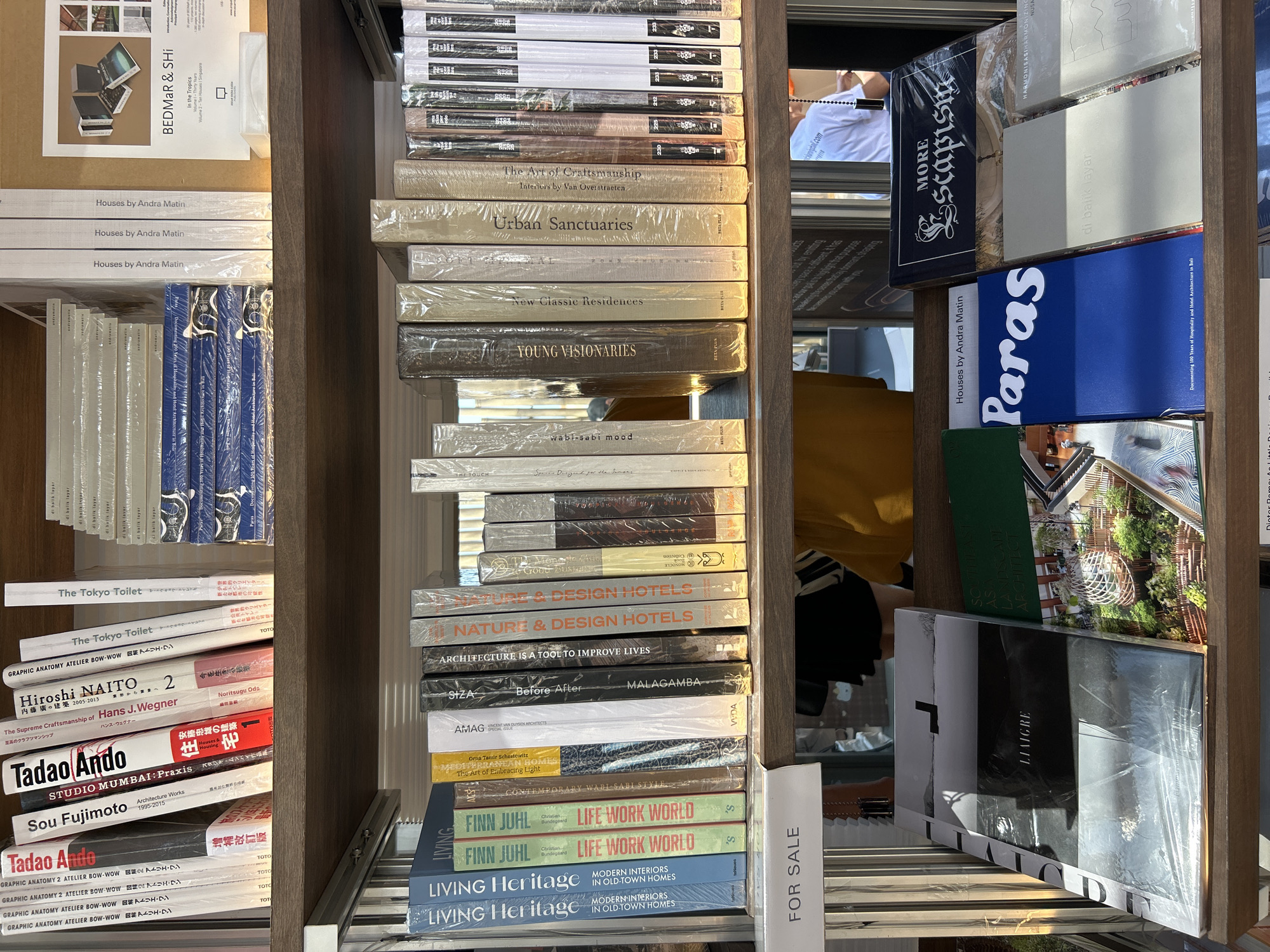 The image shows a shelf of books in a store. The books are stacked horizontally and vertically on two shelves. The top shelf displays a collection of art and design books with various titles such as "The Tokyo Toilet," "Sou Fujimoto," and "Tadao Ando." Some of these books are shrink-wrapped. To the right of the books, a poster with the text "BEDMar & SHI" is visible. The bottom shelf has a wider variety of books, including titles like "Living Heritage," "Finn Juhl Life Work World," "Architecture is a Tool to Improve Lives," and "Nature & Design Hotels." There are also some books in Indonesian language such as "di balik layar." A "FOR SALE" sign is placed on the edge of the bottom shelf. In the background, indistinct figures of people can be seen. The overall environment suggests a bookstore or an exhibition space. The books and posters suggest an architectural and design focus. The presence of Indonesian language on some books and the "di balik layar" (behind the scenes) title could hint at a location in Indonesia. Given the prominent display of books on architecture and design, it is plausible that this scene is located in Denpasar, Indonesia, a city known for its cultural and artistic offerings.