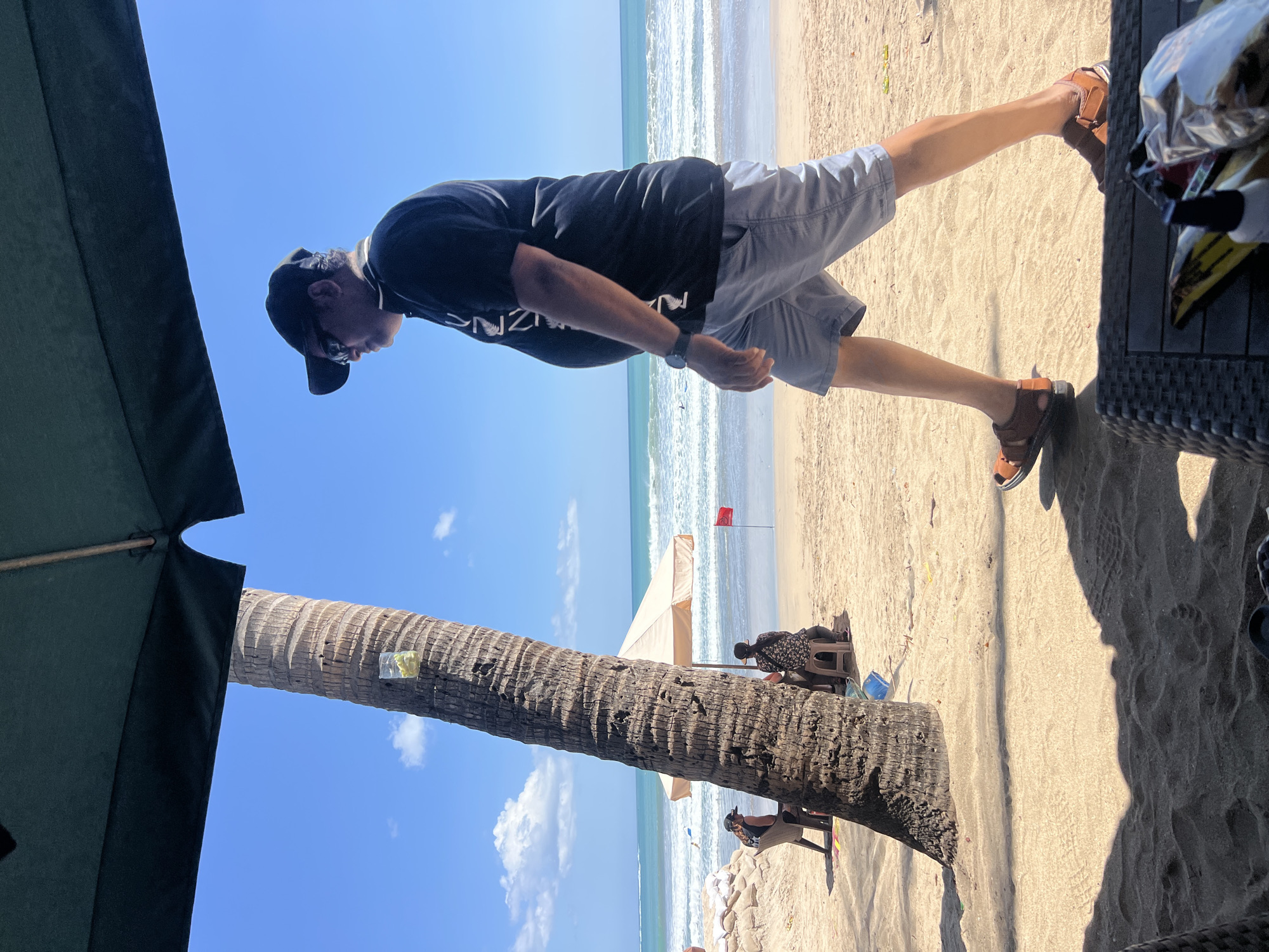 The image depicts a sunny day at Kuta Beach in Bali, Indonesia, with a man walking across the sand. He is wearing a black t-shirt, light blue shorts, a black baseball cap, sunglasses, and brown sandals. In the background, the ocean with gentle waves is visible, along with a red flag planted in the sand, possibly indicating a swimming warning. Several beach umbrellas and palm trees dot the landscape, and in the distance, a few people are seated under an umbrella. The foreground shows a sandy surface with shadows, suggesting a shaded area from which the photo was taken, and a glimpse of a dark-colored piece of furniture.