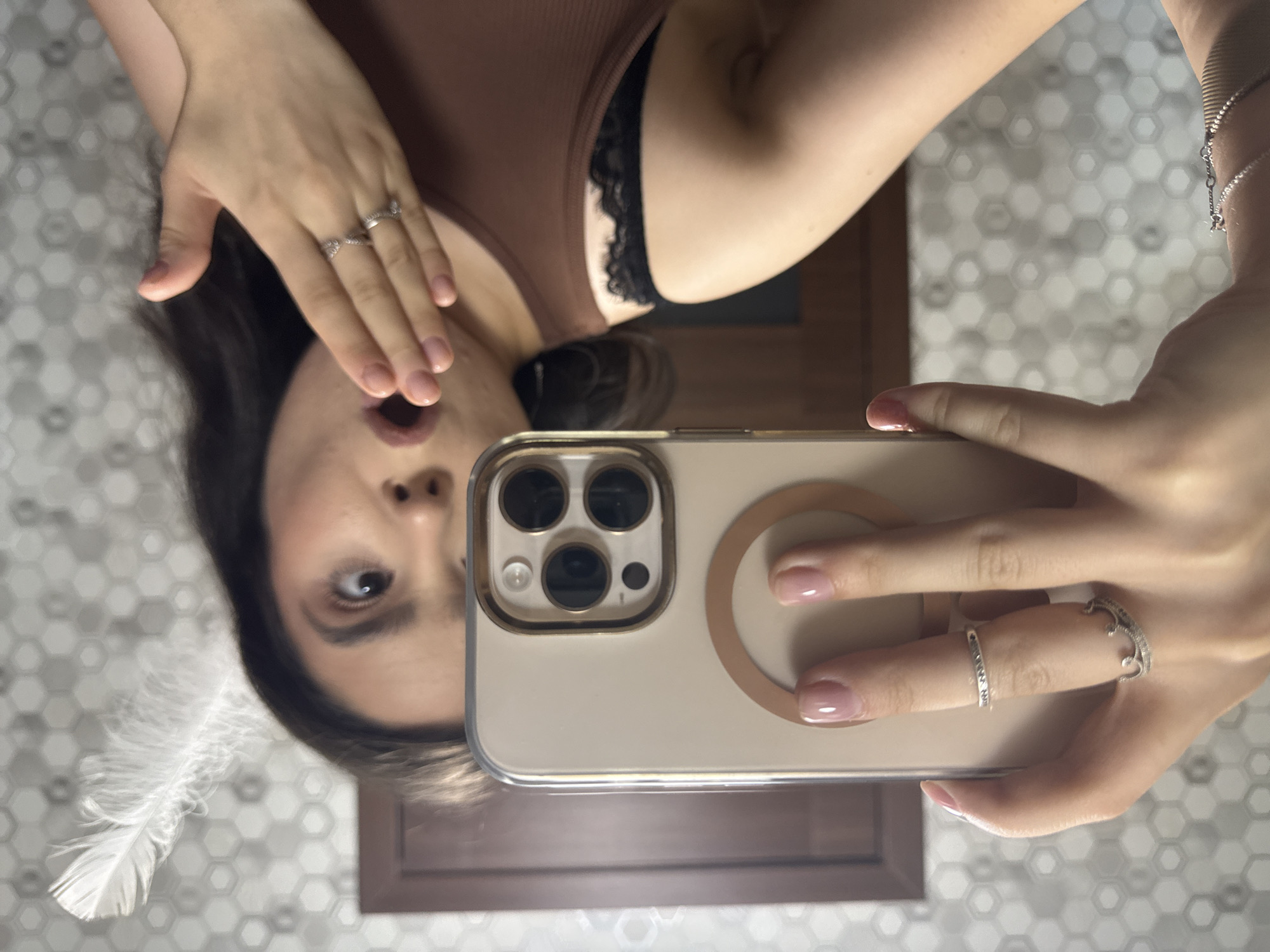 A young woman with dark hair is taking a selfie in a bathroom. She is wearing a brown tank top with black lace trim and several rings. A white feather is sticking out from behind her head. The background consists of a hexagonal tiled wall and a wooden door.
