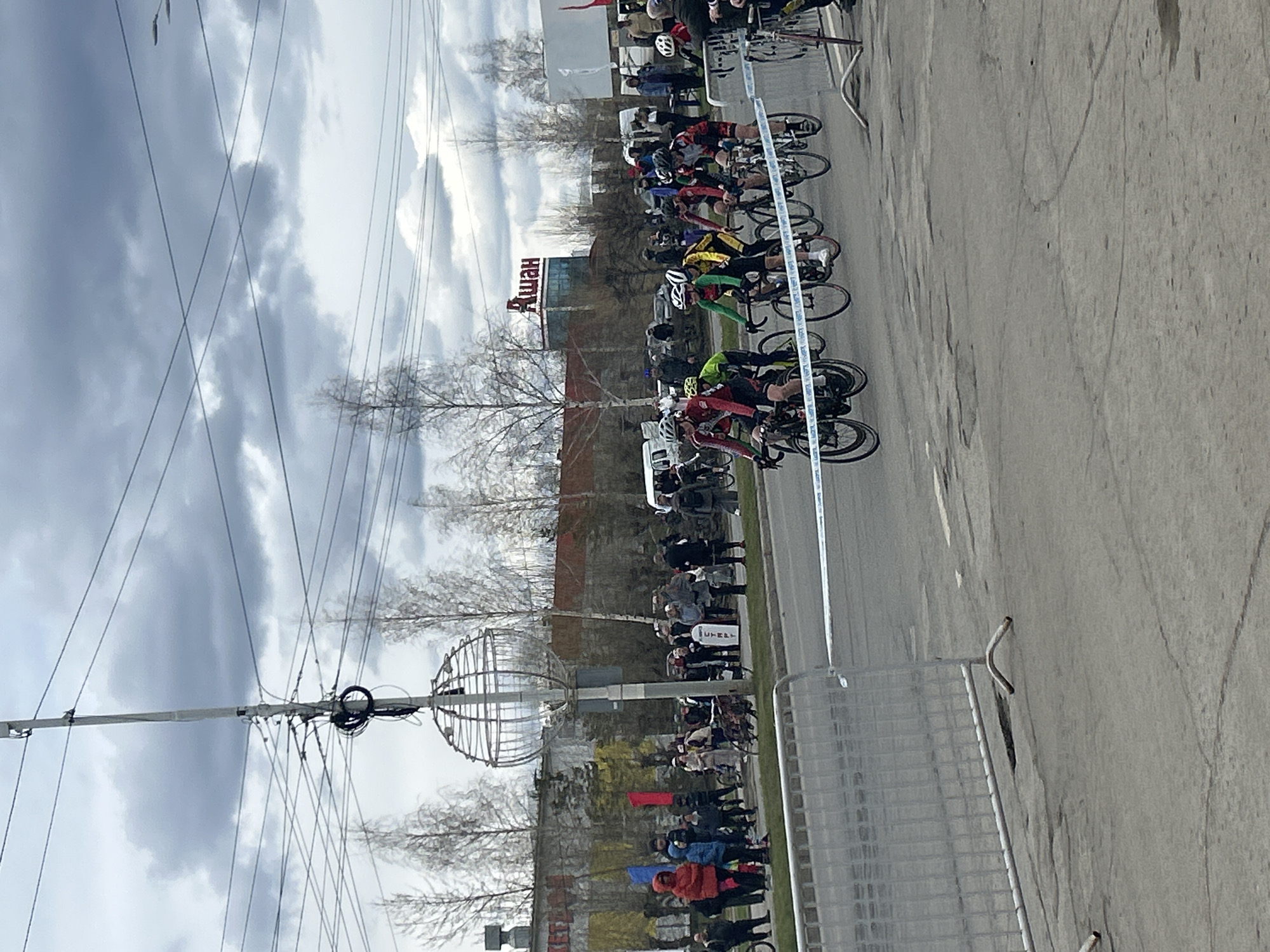 The image shows a cycling race in Izhevsk, Russia. Cyclists are racing down a street lined with spectators. A large "Ашан" (Auchan) supermarket sign is visible in the background. Power lines are visible overhead, and the sky is overcast. A metal fence is present on the side of the road, along with a spherical metal structure.