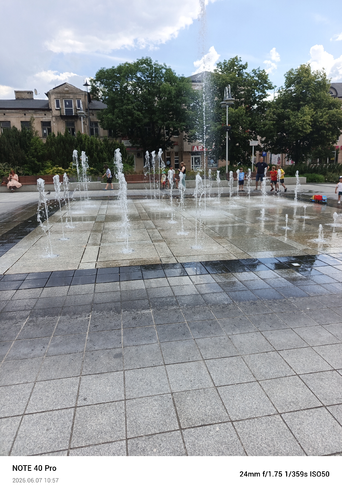 This image depicts the market square (Rynek) in Piaseczno, Poland, featuring an interactive splash pad fountain. The ground is paved with square granite tiles, primarily light grey with a darker grey band crossing the middle ground. Numerous vertical water jets rise from the pavement, creating a public fountain where children are actively playing.
In the mid-ground, a man in a dark blue shirt and shorts stands within the wet area, observing three boys. One boy in a blue shirt stands in the water, another in a yellow shirt runs left, and a third in a red shirt runs right. Further to the right, a child in a white hat is near a red and blue toy truck. On the left side of the fountain, a pigeon stands on the wet tiles. Along the left perimeter, a woman in a light-colored dress sits on a wooden slatted bench, looking down at a mobile phone. Another woman in a dark top stands on the right side of the fountain.
The background features a line of green trees and shrubs, behind which several multi-story buildings are visible. On the far left, an older, somewhat weathered building with a satellite dish on its roof and a small balcony stands. Towards the center-right, a building clearly displays an "APTEKA" (pharmacy) sign. The sky above is partly cloudy with blue patches and white cumulus clouds. The scene is brightly lit by daylight, casting soft shadows. The timestamp indicates the photo was taken on 2026.06.07 at 10:57.