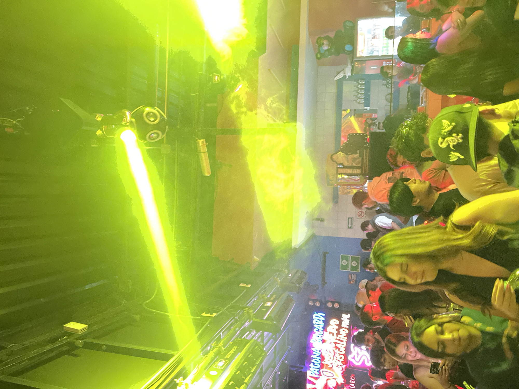 The image depicts a crowded indoor setting, likely a nightclub, in San Andrés Cholula, Mexico. A large group of people is present, some of whom are visible in detail. The illumination is intense, with a strong green light dominating the scene, emanating from stage lighting fixtures. Stage lighting equipment is also visible. A bar area is present in the background, with signage advertising "Patrona Bacardi." A sign on the wall shows Mexican symbols.