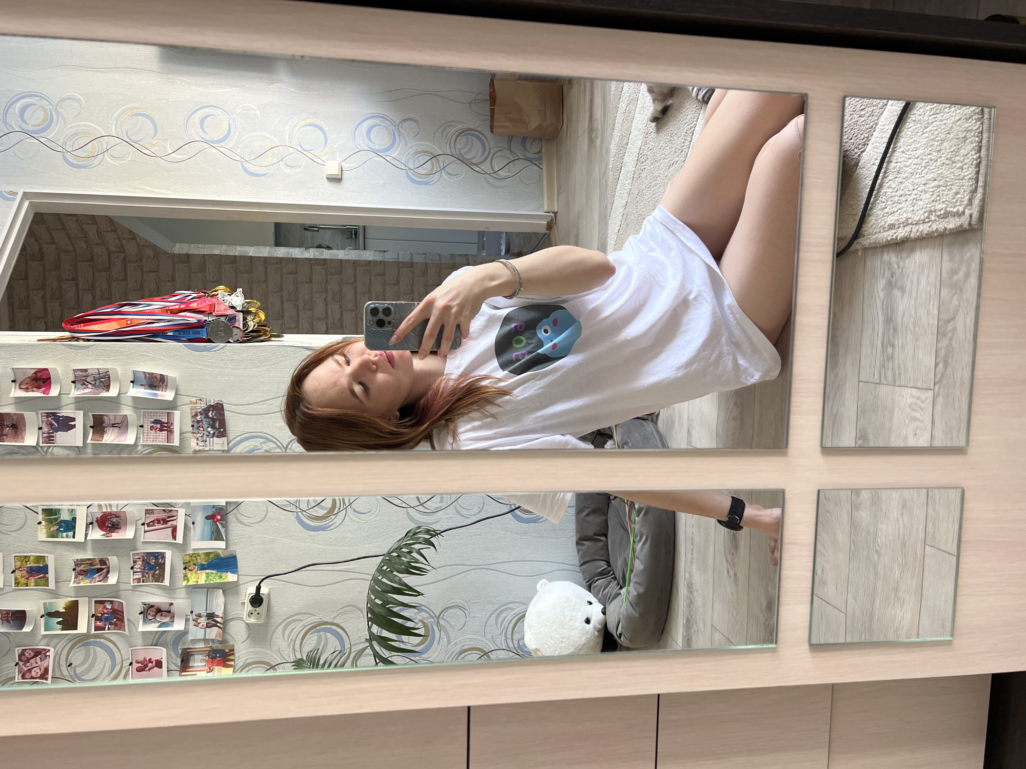 The image shows a selfie of a woman reflected in a multi-paneled mirror. The woman is lying down, holding her phone up to take the picture. She is wearing a white t-shirt.
Behind the woman, the reflection shows a room with patterned wallpaper. On a shelf, there are medals and photographs. There is also a stuffed polar bear toy and a houseplant. The location may be an apartment in the city of Chaikovsky, Russia based on visual elements within the reflections.