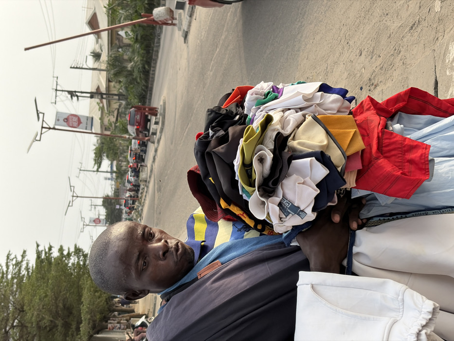 An individual carries a large bundle of colorful clothing on their shoulder, likely for sale or transport. The man has dark skin, a shaved head, and wears a collared shirt and jacket. The background features a street scene with cars, streetlights, trees, and a billboard in the distance, suggesting an urban setting. The photograph appears to be taken from a close perspective, emphasizing the weight and size of the clothing bundle.