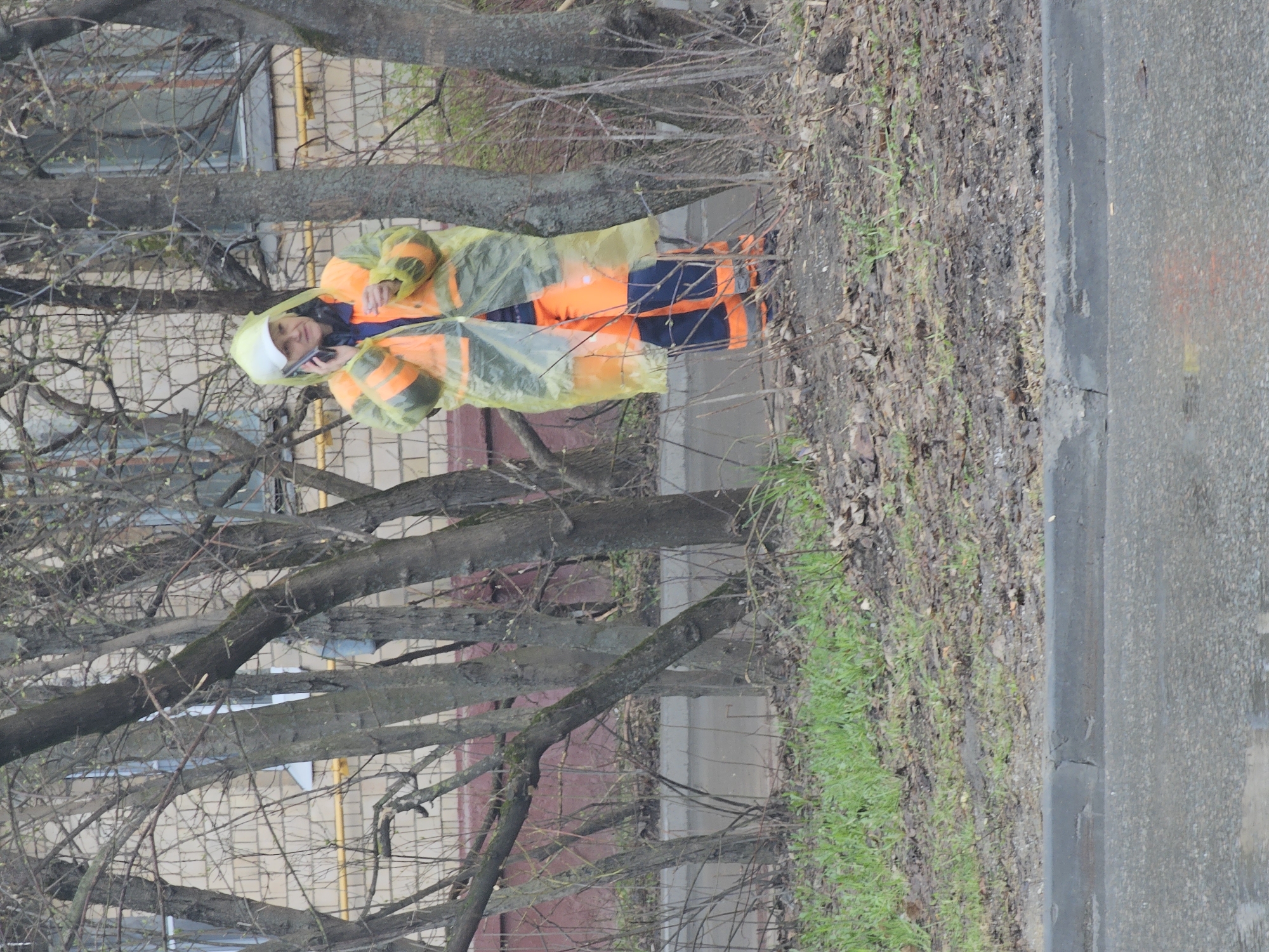 A street scene in Khimki, Russia, depicts a person dressed in an orange safety vest and a transparent yellow rain poncho. The individual is standing near a sidewalk, talking on a mobile phone. Bare tree branches fill the upper left portion of the frame. A brick building with light-colored rectangular brickwork is partially visible behind the trees. To the right of the sidewalk is a patch of ground covered in dried leaves and sparse grass. A gray asphalt road runs along the right edge of the image.