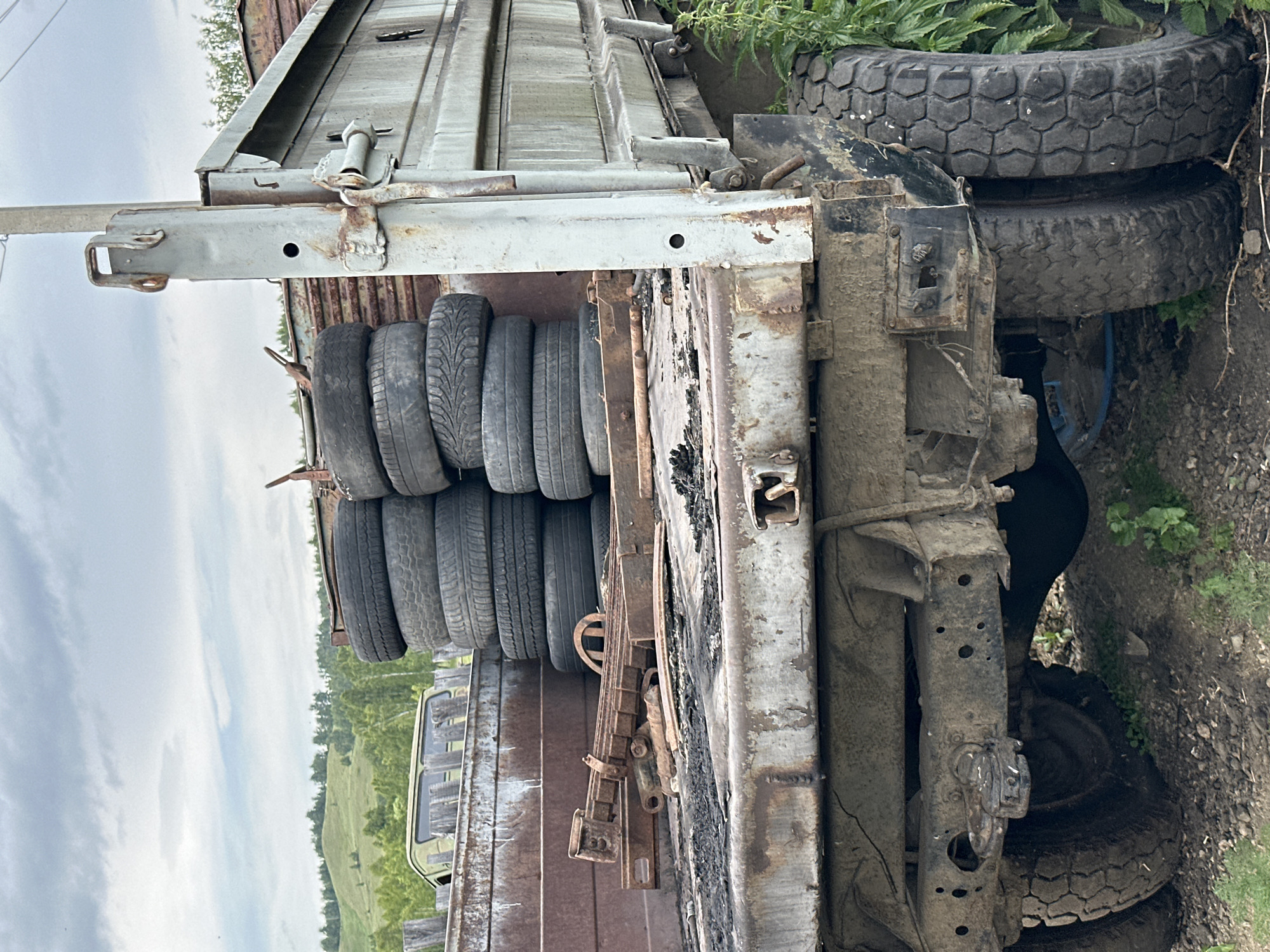 An exterior, medium shot captures the rear of a dilapidated, flatbed truck loaded with stacks of tires. The truck, which appears rusted and worn, is parked outdoors on a gravel and dirt surface. In the background, rolling hills covered in green trees and vegetation rise under a cloudy sky. The truck bed is filled with numerous tires, some stacked vertically, and also contains some corroded metal parts. The truck's chassis and tires are visible, showing signs of age and wear. The scene suggests a rural or industrial setting where old vehicles and materials are stored.
