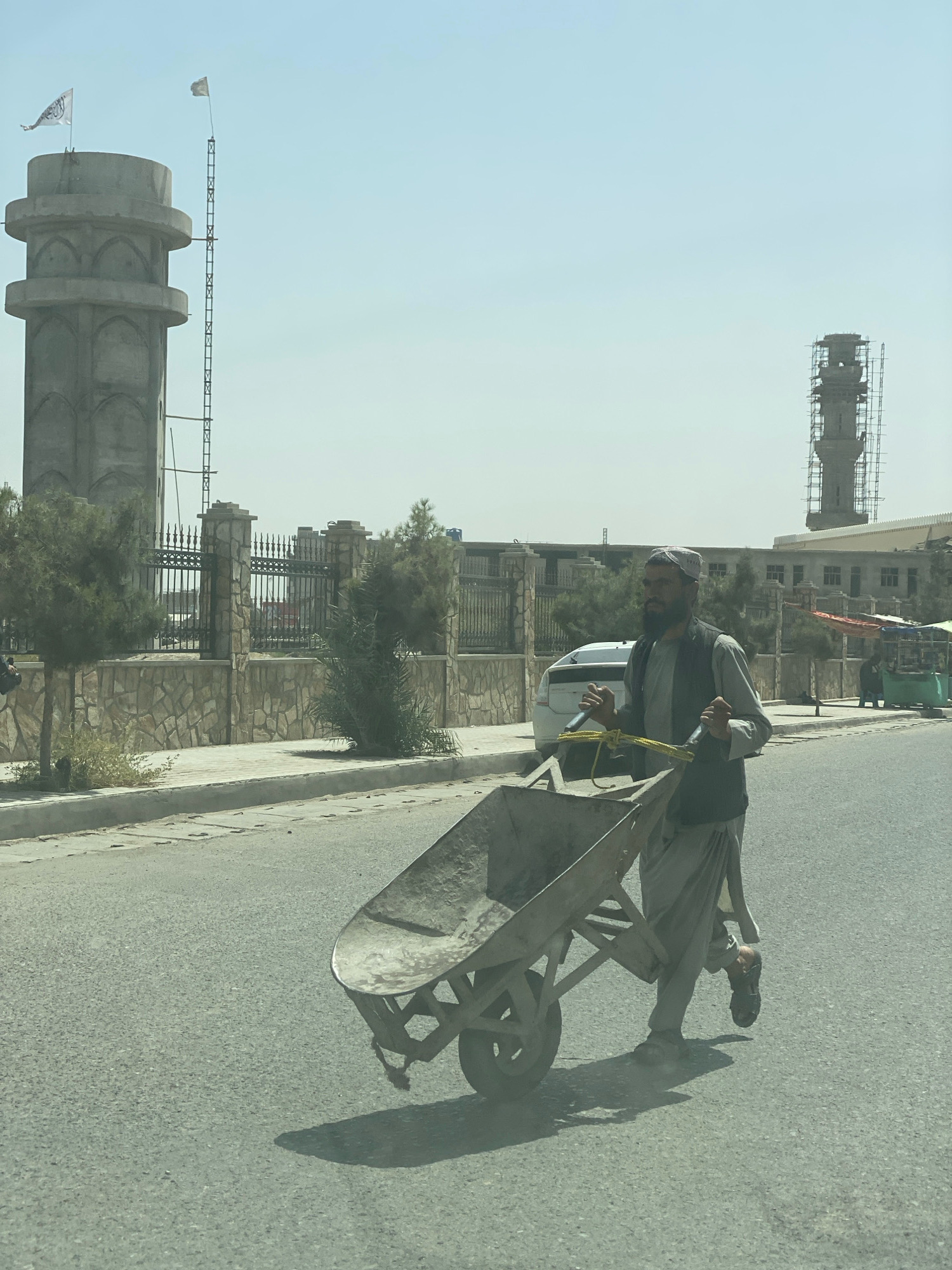 A man is pushing a wheelbarrow down a paved road in Kandahar, Afghanistan. He is wearing a light gray traditional outfit and a white cap. He has a dark beard and is looking forward. The wheelbarrow is made of metal and has a yellow rope tied to it. In the background, there is a large stone building with a tall tower and a fence. There are also trees and a blue sky. The sun is shining, casting a shadow from the man and the wheelbarrow. The scene appears to be during daylight.