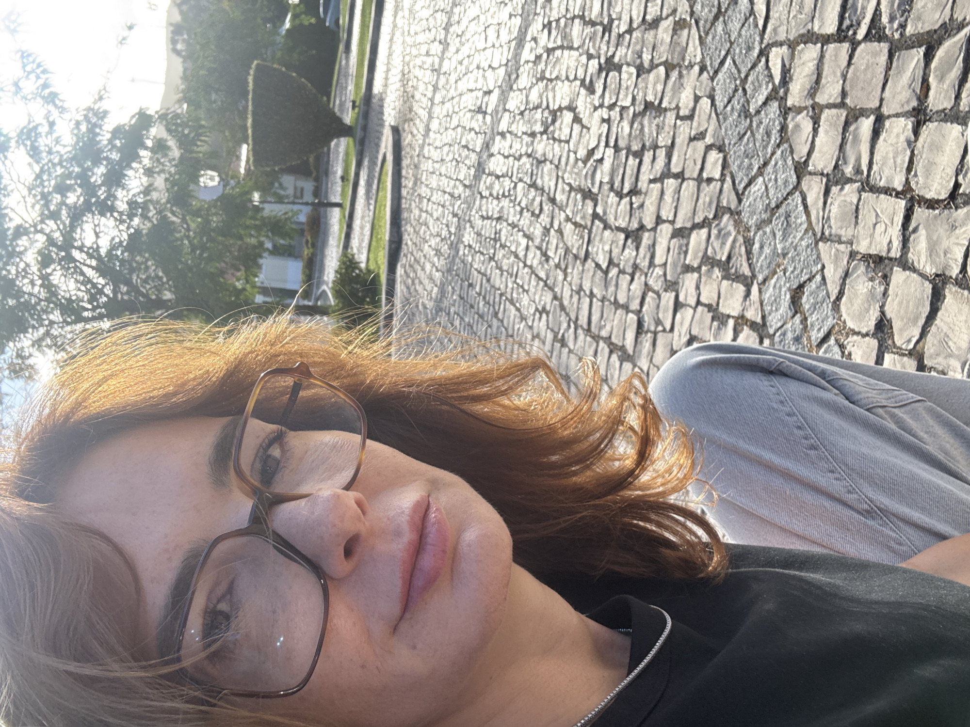 A woman with shoulder-length, reddish-brown hair and freckles is captured in a close-up selfie. She is wearing large, brown-rimmed glasses. Her lips are slightly parted, and she appears to be looking towards the camera. She is wearing a black t-shirt with a thin silver necklace and light blue jeans. The background is an outdoor scene with a cobblestone pathway, green grass, and trees. A street lamp is visible in the distance, and the sun is shining, casting light on the scene. This scene is located on Rua General Luís do Rego in Torres Vedras, Portugal.