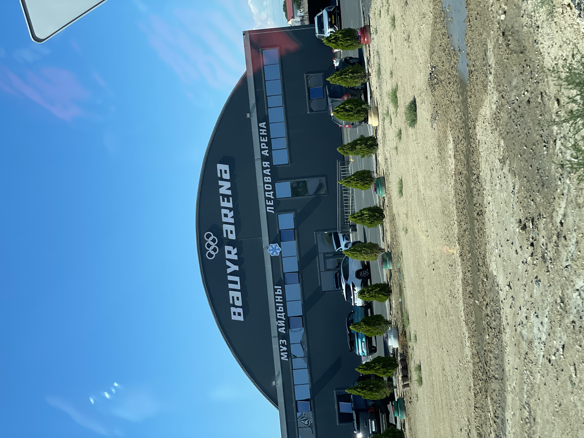 The image captures the "Baryr Arena" in Aktau, Kazakhstan, under a clear blue sky. The arena is a large, dark-colored building with a curved roof, prominently displaying the Olympic rings logo and the text "BAUYR ARENA" in white. Below this, in smaller white lettering, is the Russian phrase "ЛЕДОВАЯ АРЕНА," which translates to "Ice Arena," and the Kazakh phrase "МУЗ АЙДЫНЫ," also meaning "Ice Arena." Several rows of dark-paneled windows line the facade.
In front of the arena, a paved area serves as a parking lot, where several cars are visible. These include a dark SUV on the left, a turquoise SUV, a white SUV, and a darker SUV parked further down. A row of small, manicured evergreen trees stands between the parking lot and a sandy, somewhat uneven ground in the foreground. Some of these trees are planted in what appear to be repurposed tire planters painted in various colors. The ground in the foreground shows signs of recent rainfall or drainage, with muddy patches and shallow pools of water. The overall scene depicts an outdoor view of a sports facility.
