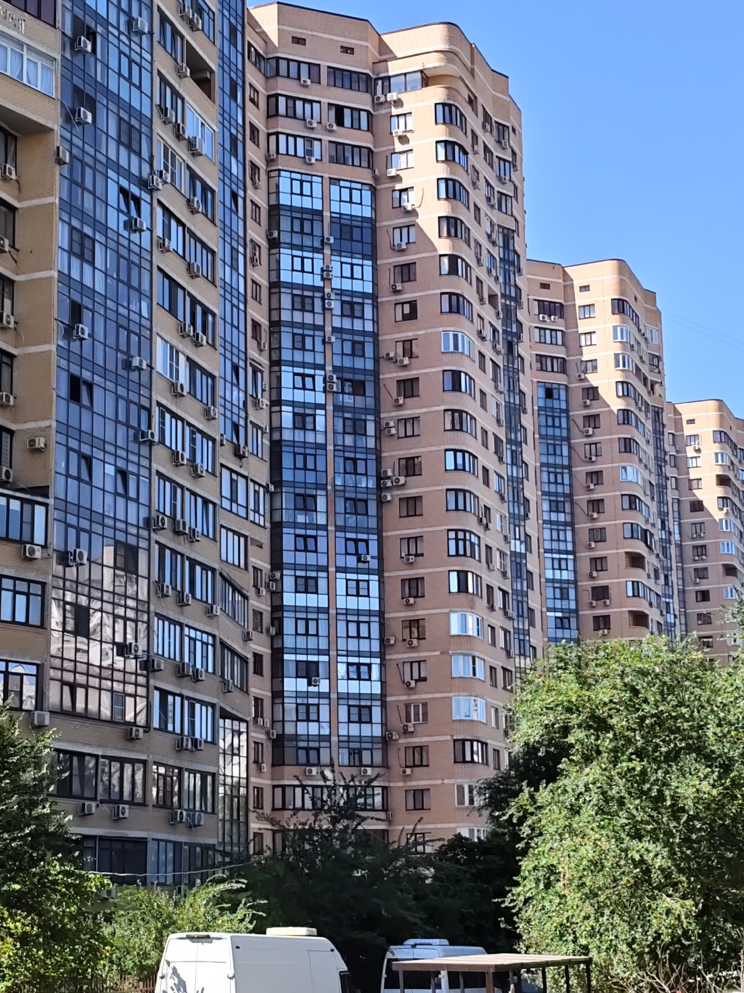 The image shows a street view of several tall residential buildings under a clear blue sky. The buildings are modern, with balconies and many windows. The facades are primarily beige brick with some sections of dark blue glass. Numerous air conditioning units are visible mounted on the exterior walls of the buildings. In the foreground, dense green trees partially obscure the lower portions of the buildings. A white van is parked at the bottom of the image, and part of a wooden structure, possibly a table, is also visible. The scene appears to be a typical urban residential area.