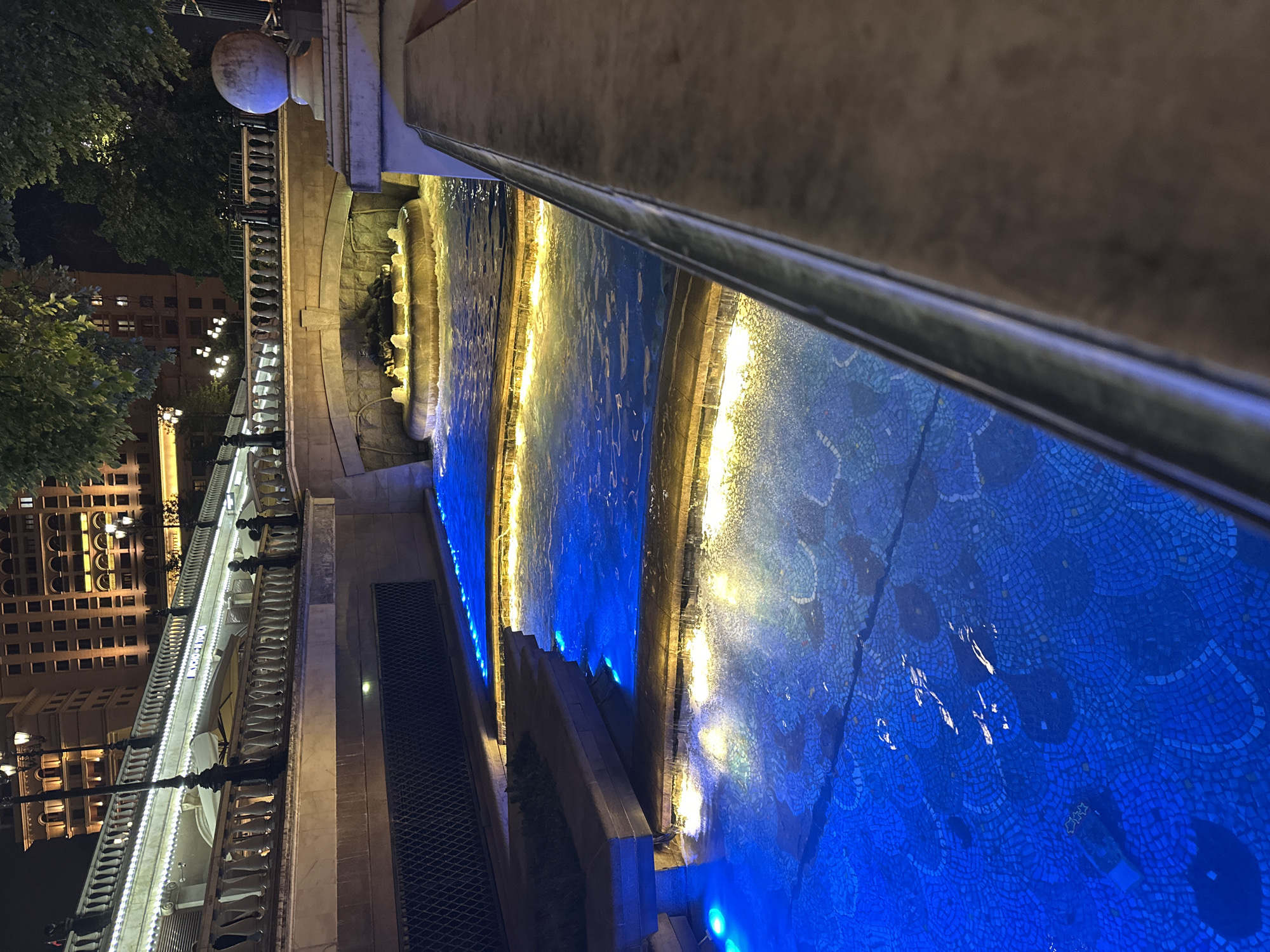 The image, rotated to its correct orientation, captures a vibrant urban night scene in Moscow, Russia. The central element is a multi-tiered water feature or canal, beautifully illuminated. The water glows a deep, rich blue from submerged lights, while the edges of each tier are lined with warm, golden-yellow lights, creating a striking visual contrast and reflections on the water's surface. The bottom of the water feature reveals intricate blue mosaic tile work, some patterns resembling scales or abstract designs. Small sculpted figures or elements can be discerned within the fountain's structure, particularly where the water flows from one tier to the next.
To the left of the water feature, an ornate stone balustrade, likely part of a bridge or promenade, runs parallel to the illuminated canal. This balustrade is brightly lit by white string lights, accentuating its architectural details. On a sign attached to the balustrade, the word "МОСКВА" (Moscow) is clearly visible, confirming the location. Beyond the balustrade, several multi-story buildings with numerous illuminated windows rise against the dark night sky, partially framed by dark green foliage. Ornate streetlights with multiple globes are also visible along the promenade. A dark, possibly metallic, grate or pathway is situated alongside the water feature on the far left. The right side of the foreground is occupied by a dark, smooth wall or railing, from which the photograph appears to have been taken. The time of day is unequivocally night, as indicated by the dark sky and the reliance on artificial lighting. The scene conveys a tranquil yet visually rich urban atmosphere.