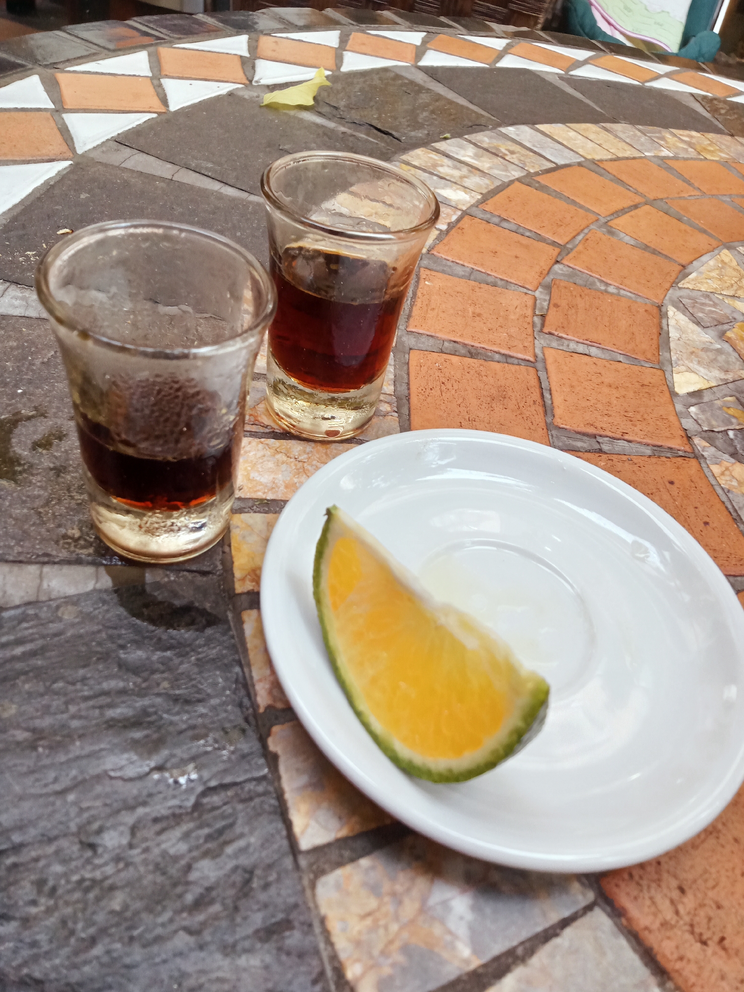 The image captures a close-up, slightly elevated view of two shot glasses and a white saucer with an orange slice, resting on a mosaic-tiled table. The setting appears to be an outdoor or semi-outdoor cafe or restaurant in Nha Trang, Vietnam.
The main subjects are two small, clear glass shot glasses, each containing a dark, reddish-brown liquid, likely coffee or a dark spirit. The glass on the left is closer to the viewer and appears to have less liquid than the one on the right. Both glasses show some condensation or residue on their exteriors, suggesting a cool beverage or humid environment.
Adjacent to the glasses, a small, round white ceramic saucer holds a wedge of orange. The orange slice is vibrant, with bright orange flesh and a distinct green rind, indicating it might be a type of sour orange or lime often used as a garnish.
The table features an intricate mosaic pattern, consisting of irregularly shaped tiles in various earthy tones, including terracotta orange, white, and different shades of grey and beige. Some areas of the table are covered in larger, darker grey, slate-like tiles. A small, yellow, possibly dried leaf is visible on the table surface in the background.
The lighting is soft and diffused, suggesting it could be daytime, possibly under an awning or on an overcast day, as there are no strong shadows. The overall scene conveys a casual, relaxed atmosphere, typical of a beverage serving in a tropical location. There is no visible text in the image.