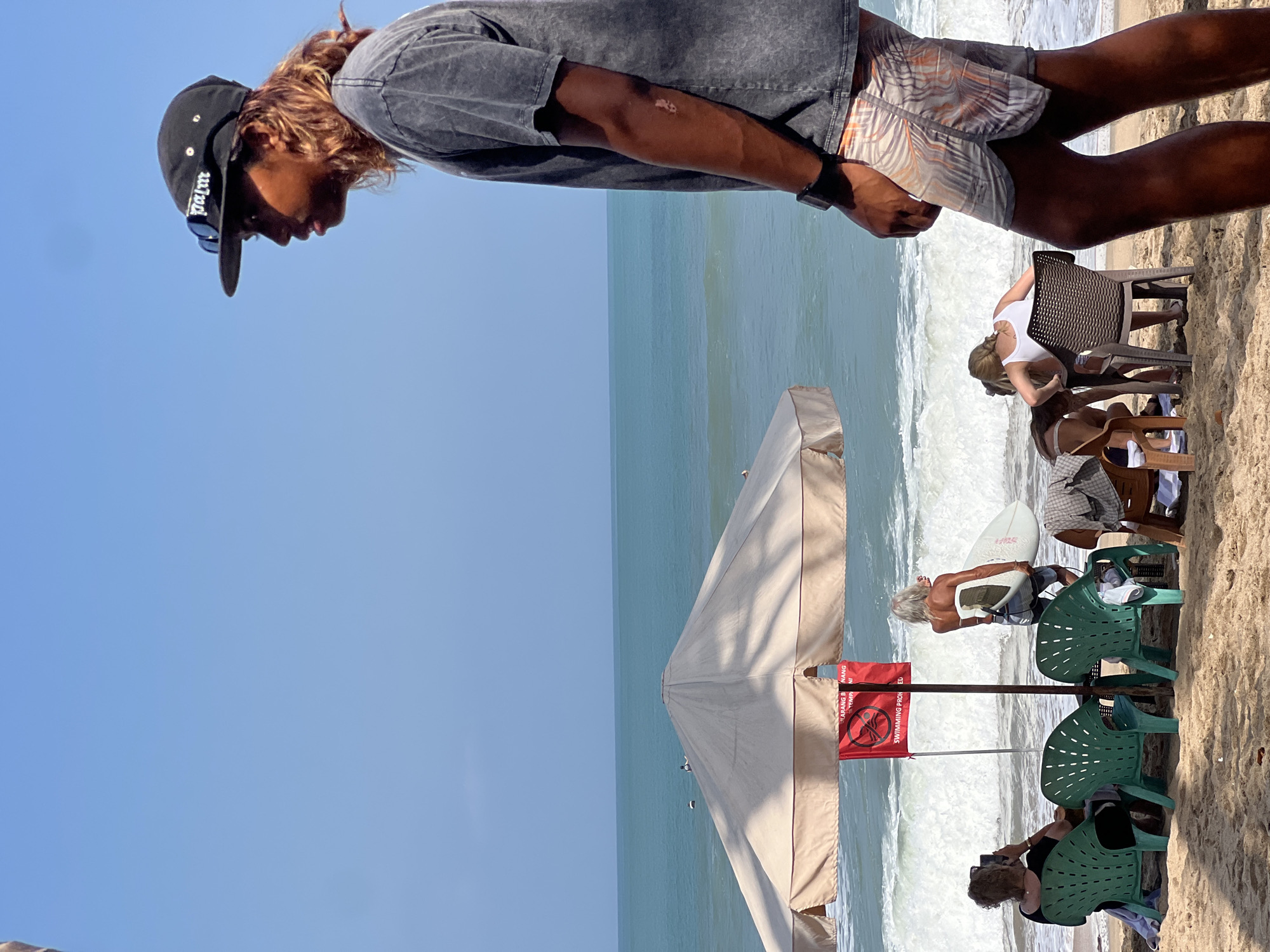 In Kuta, Indonesia, a beach scene unfolds under a clear blue sky. In the foreground, a young man wearing a black baseball cap with "palm" inscribed on it, sunglasses, a dark gray t-shirt, and patterned swim shorts stands in profile, facing right. His right arm, adorned with a black watch, is visible, showing a scar on his forearm.
Further back, under a large, light-colored beach umbrella, several individuals are seated. On the left, a woman with curly brown hair is seated in a green plastic chair, holding a phone and taking a picture. Next to her are two more green plastic chairs. To the right of the umbrella, a woman with long, white hair stands holding a surfboard, facing the ocean. Behind her, a red sign with white lettering and a graphic of a crossed-out swimmer is attached to a pole.
In the mid-ground, two people are seated at a table with chairs. One, wearing a white tank top, is leaning forward. Another person, partially obscured, is also visible. The ocean, with moderate waves, stretches to the horizon. A few surfers are visible in the distance. The sand of the beach is light brown.