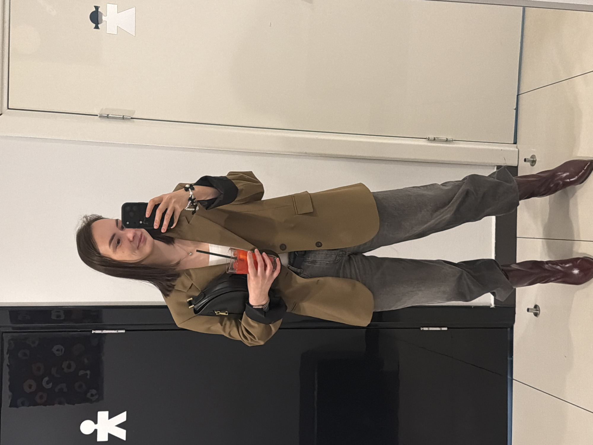 A young woman is taking a selfie in a restroom in what appears to be the city of Chaikovsky, Russia. She is wearing a tan blazer, gray pants, and brown boots, holding a drink with a straw and a black purse. The restroom has doors with gender symbols on them. The walls are white tiled.