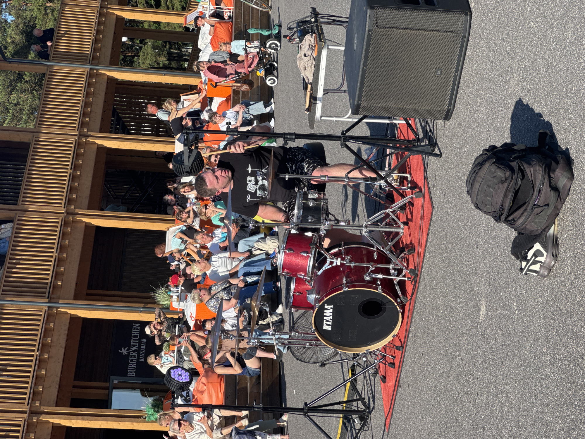 The image depicts an outdoor music performance at a venue resembling a beach bar or outdoor restaurant in Tallinn, Estonia, specifically at Rannabaar, as indicated by the signage. The foreground is dominated by a red carpet laid out on asphalt, on which a drum kit is set up. A male drummer, wearing a black t-shirt with a drum set graphic and patterned shorts, is actively playing the drums, holding drumsticks in his hands. Adjacent to the drum kit is a large black speaker cabinet. Behind the drummer and to the left, a small fan is visible. In the background, an elevated wooden structure with railings and staircases forms the main building of the venue. A crowd of people, diverse in age, are seated on wooden steps and lounge chairs, observing the performance. Among the spectators, a woman pushes a stroller with a young child. The environment appears to be a sunny day, with ample natural light casting shadows. A black backpack is placed on the asphalt in the lower right quadrant of the image. The overall scene conveys a casual, open-air entertainment event.