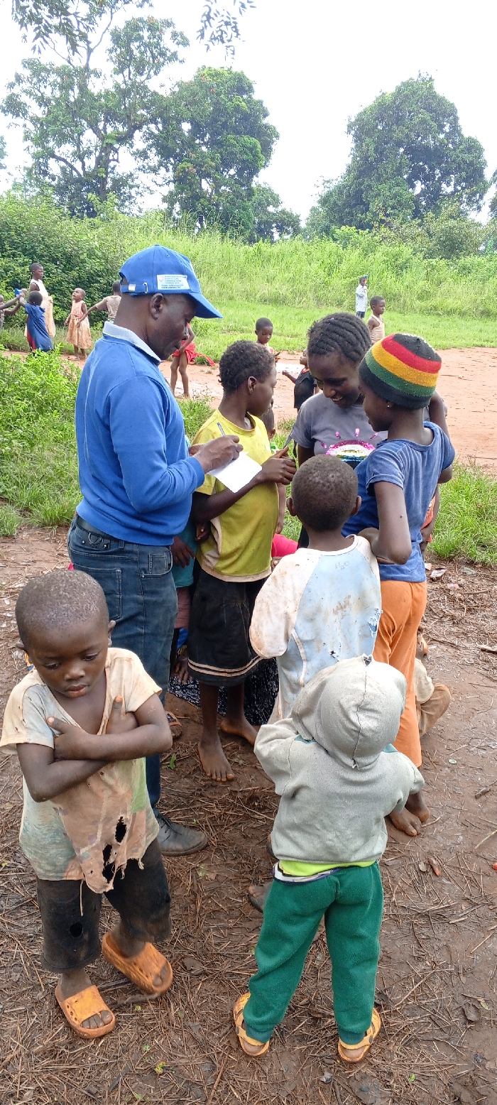 In Ngalula, Democratic Republic of the Congo, an outdoor scene depicts an adult male interacting with several children on a reddish-brown dirt ground with sparse vegetation. The adult male, with a dark complexion, wears a blue long-sleeved shirt, dark blue jeans, and a blue baseball cap featuring a white logo and text. He holds a white pad of paper and a yellow pen, appearing to write or take notes, looking downwards at the paper.
Around him, a group of children are gathered. To his right, a child in a dirty yellow t-shirt and dark shorts stands barefoot, looking towards the paper. Behind this child, another child with dark braided hair wears a dirty grey t-shirt with a colorful graphic and appears to be smiling or laughing. Adjacent to them, a child in a blue t-shirt, orange pants, and a colorful striped knit cap (red, yellow, green, black) also looks towards the paper. Another child, facing away, wears a light blue and white t-shirt that appears dirty and torn. In the immediate foreground, a small child in a light grey hooded sweatshirt and green pants, wearing light brown sandals, stands with their back to the camera. To the left foreground, a child with arms crossed wears a dirty, ripped beige/white t-shirt and dark pants, with orange slip-on sandals.
In the mid-ground and background, other children are visible, dressed in various simple, often worn garments. The environment features lush green foliage, tall grasses, and large trees against an overcast or bright sky, indicating a natural, unpaved setting. The overall scene suggests a community interaction or documentation event involving children in a rural or semi-rural location.