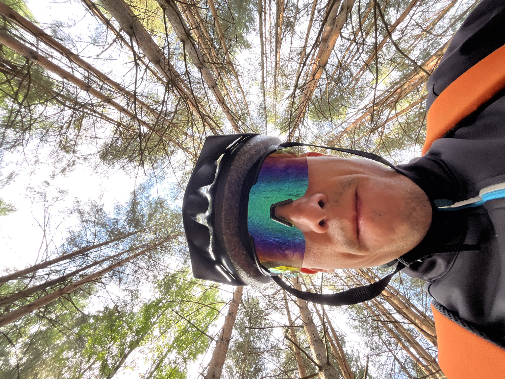 The image shows a first-person perspective of a person wearing a black cycling helmet and multi-colored mirrored cycling glasses. The person is looking directly at the camera. They are wearing a black cycling jacket with blue and white accents and an orange backpack strap visible over their left shoulder. The background consists of a dense canopy of pine trees, with the sky visible through the branches. The camera angle is looking upwards into the trees. The location is indicated as Chaikovsky, Russia.