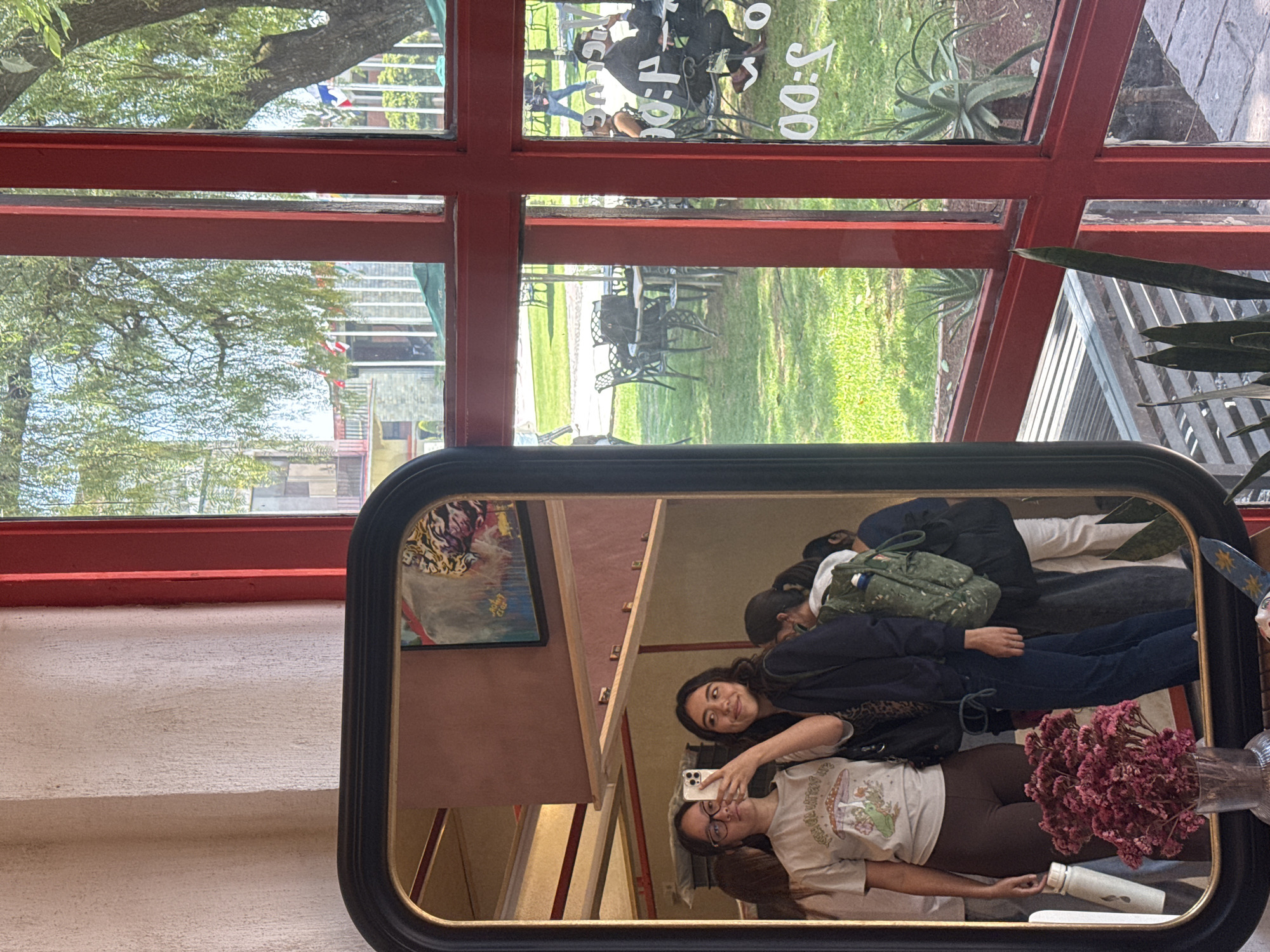 The image captures a mirror reflection of two young women taking a selfie. One woman is wearing glasses, a T-shirt with a mushroom design, and brown leggings, while the other wears a dark jacket. They hold a phone to capture the picture. The mirror is framed in black and gold.
The mirror sits against a white wall within a building featuring a large red-framed window. The window looks out onto a grassy area with trees and outdoor furniture, suggesting a patio or garden setting. Through the window is visible the cityscape of San Andrés Cholula, Mexico. In the reflections on the window, the reflection of “Club 3:00” can be seen.