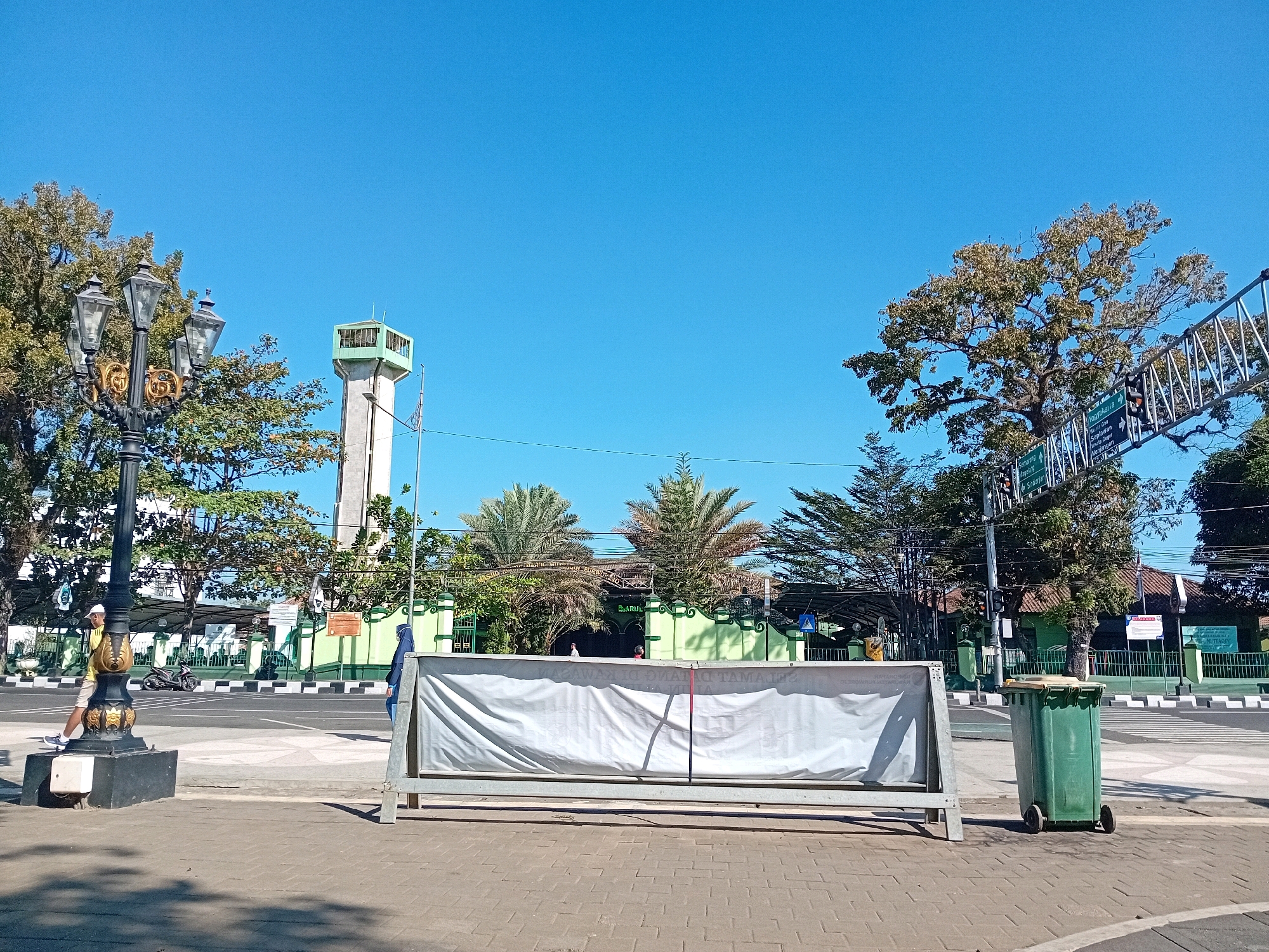 The image shows a street scene in Purworejo, Indonesia, on a clear, sunny day. In the foreground, a large white banner is erected on a metal frame, obscuring the view of a building behind it. To the right of the banner is a green trash can on wheels. A paved sidewalk with brick patterns is visible in the immediate foreground.
Further back, across a street with pedestrian crossings and black and white striped curbs, is a green building with a prominent minaret tower to its left. Trees with green foliage are scattered throughout the scene. A decorative street lamp with multiple globes stands to the left of the banner. A person wearing a yellow shirt and white shorts is walking on the sidewalk near the street lamp. Another person, wearing a blue headscarf, is standing near the banner on the right side of the image. In the middle ground, a motorcycle is parked on the side of the road. A metal structure with signage is suspended over the road in the upper right quadrant of the image. The sky is a clear, bright blue.