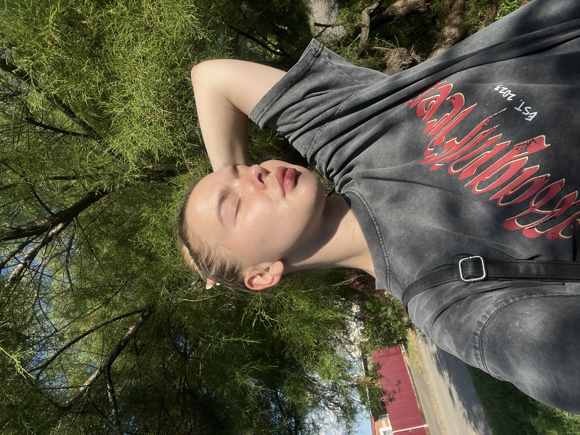 The media file is a selfie taken by a young woman outdoors, likely in Tikhoretsk, Russia. She is lying down, with her head resting on her arm, eyes closed, and a serene expression on her face, suggesting relaxation or a moment of peace. The setting is a lush green environment with trees and foliage creating a natural backdrop. Sunlight filters through the leaves, casting dappled shadows and illuminating her skin.
She is wearing a dark gray, acid-washed t-shirt with a bold red graphic print and the text "EST. 2023" printed in white. A black strap, possibly from a bag, is visible across her chest. Her ear, adorned with a small hoop earring, is also visible. The background shows hints of a red building and a paved area, possibly a street or path, suggesting an urban or suburban park area.
The time of day appears to be daytime, given the bright sunlight. The overall mood conveyed is one of tranquility and enjoyment of the outdoors.