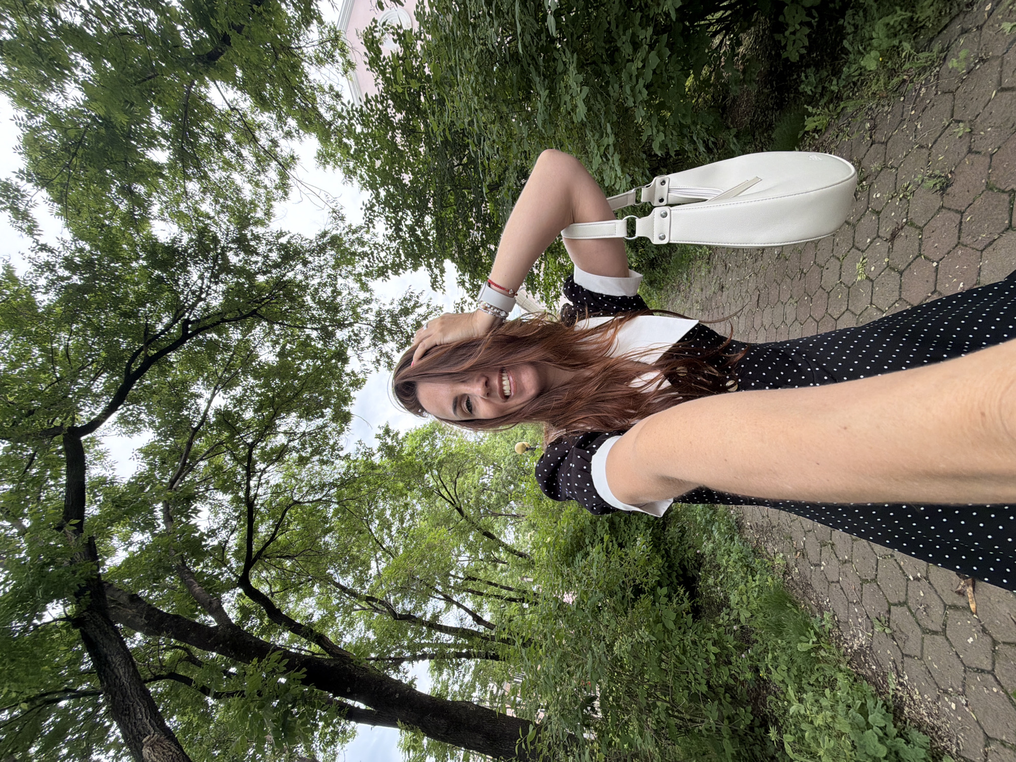 The image depicts a woman taking a selfie outdoors. She has long, reddish-brown hair and is smiling, with her right arm extended towards the camera. She is wearing a black and white polka dot dress with a white collar and cuff. She is also carrying a white handbag. The background shows a park-like setting with trees and foliage. A paved path with hexagonal stones is visible. There is a light pink building visible in the distance through the trees. The overall scene suggests a casual outing in a city park. The specific location in Birobidzhan, Russia cannot be determined from the visual information alone.
