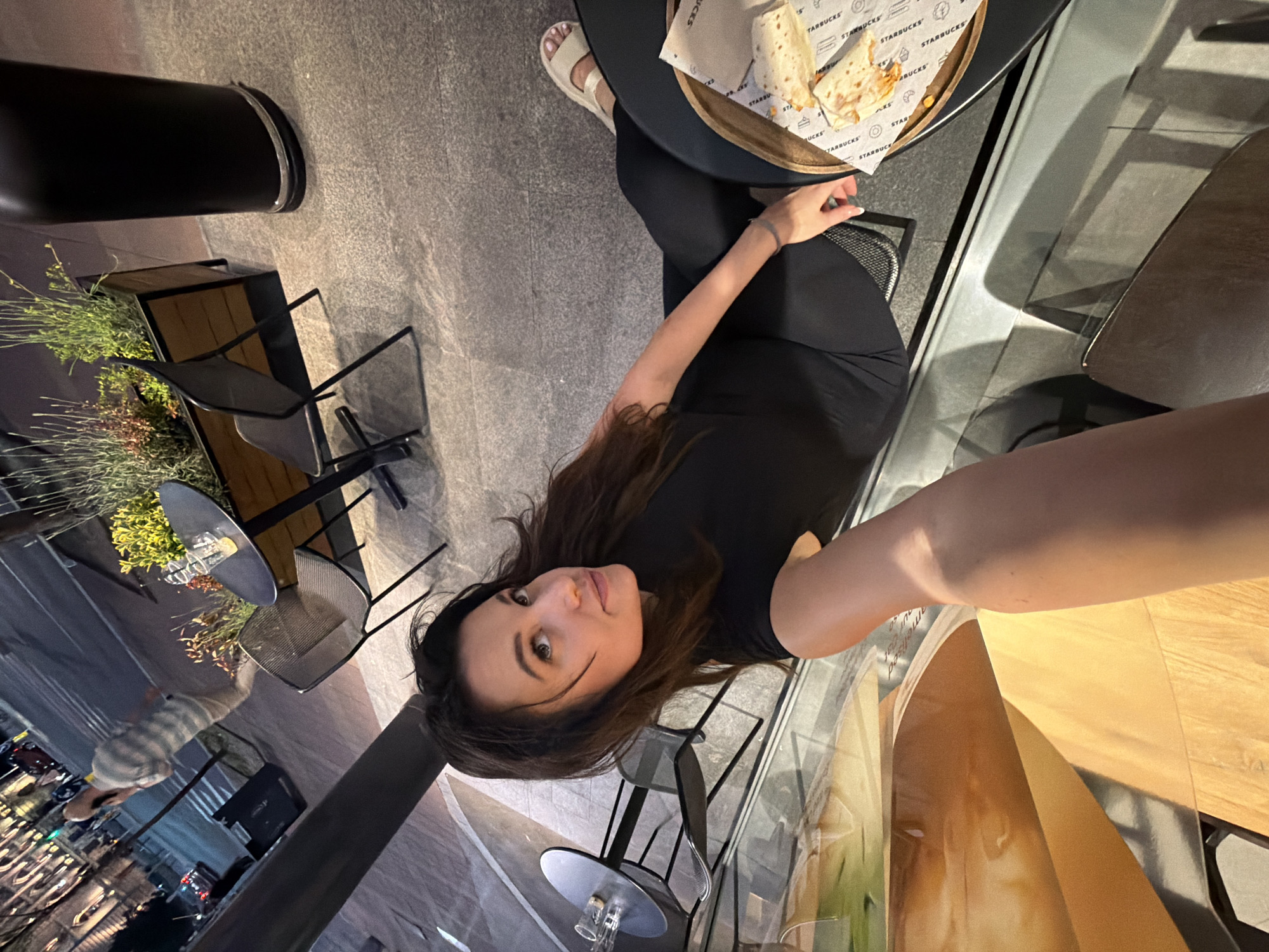 A woman is sitting at an outdoor cafe table, likely taking a selfie. She is wearing a black t-shirt and black pants, and her brown hair is loose. On the table in front of her is a plate of food, likely from Starbucks, and she is wearing sandals. The cafe has black metal chairs and tables, and there are plants nearby. Through the glass partition, one can see the city street, suggesting an urban environment. The presence of the Serbian language logo and the city landscape leads me to assume that she is in the Vracar Urban Municipality, Serbia.