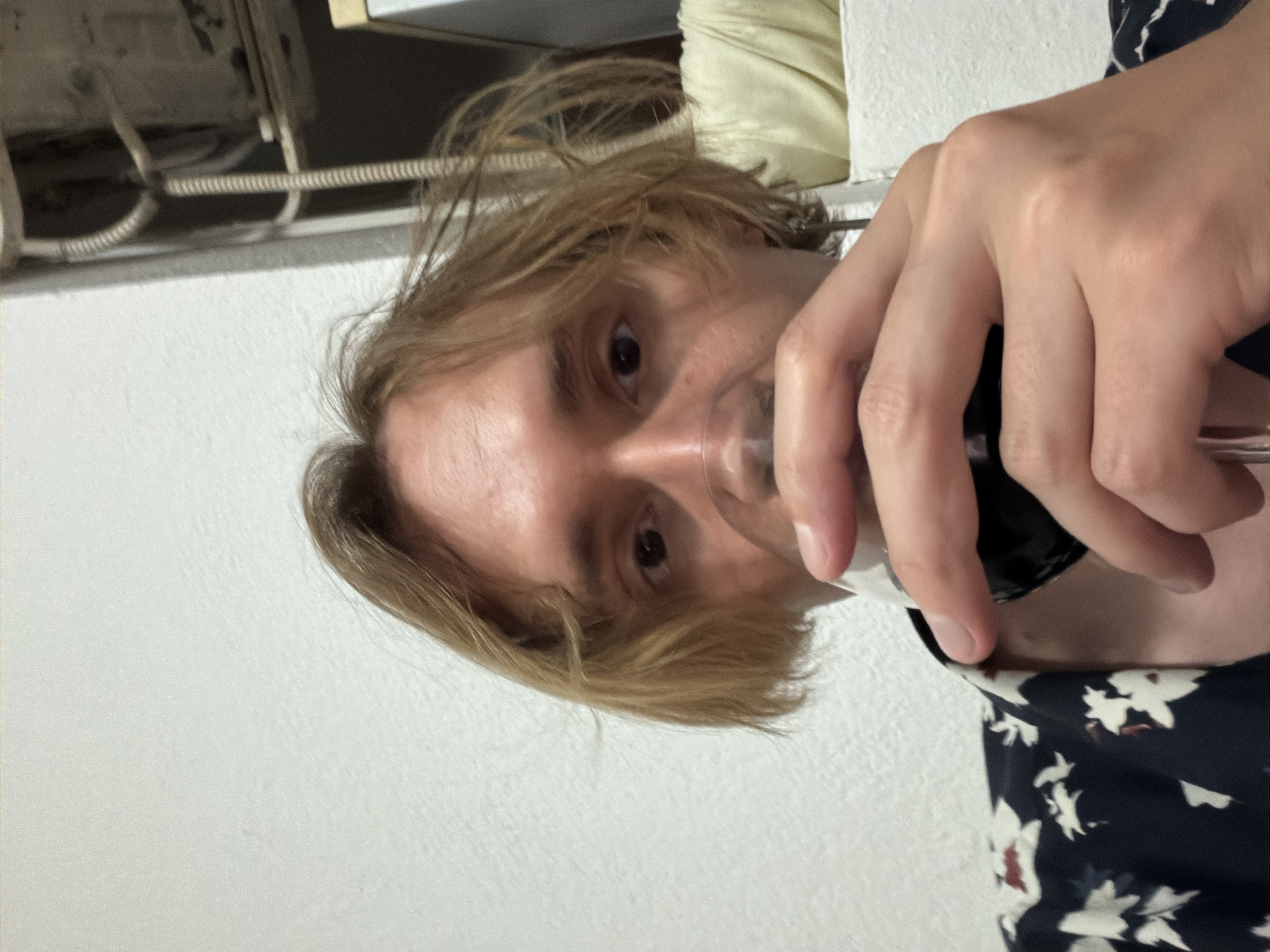 A person with light brown, shoulder-length hair is holding a wine glass in their right hand, near their face. They are wearing a floral print, dark blue top. The background is blurred but appears to be an interior setting with a white wall and some visible pipes near the ceiling. The location in the city of Budva, Montenegro, cannot be determined from the image.
