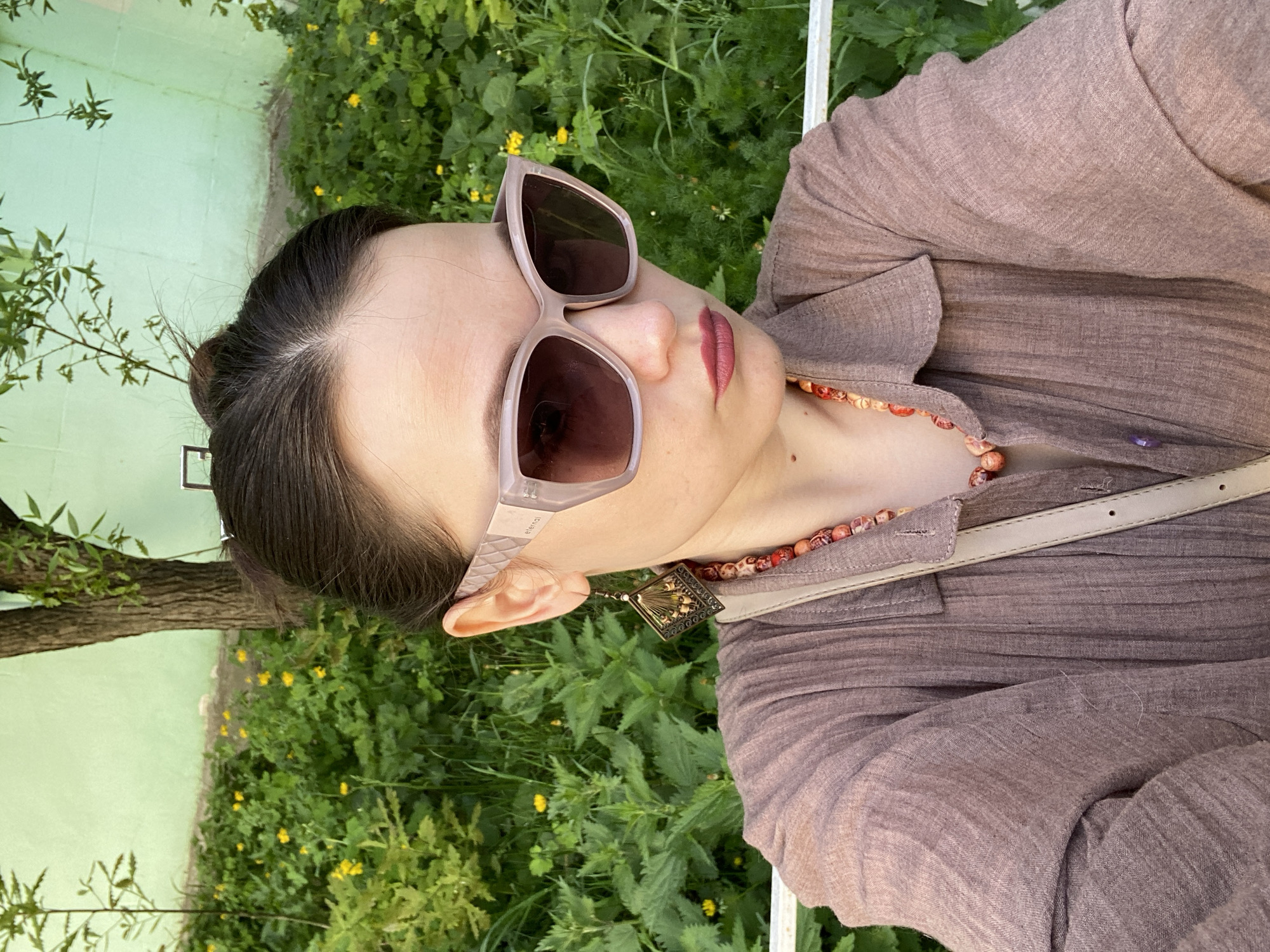 A woman wearing sunglasses and a light brown button-down shirt with a necklace and pendant is posing for a photo outside in Chaikovsky, Russia. The photo is taken at an angle, showing the woman's face, neck, and upper chest. She has dark hair pulled back in a bun and fair skin. The sunglasses have a light-colored frame and dark lenses. The necklace consists of red and orange beaded stones. The pendant appears to be bronze in color with an elaborate design. A light-colored strap from a bag or purse is visible across her chest. The background includes green foliage, yellow flowers, and a tree trunk. A painted green wall can also be seen. The environment seems to be outdoors in a park or garden setting.