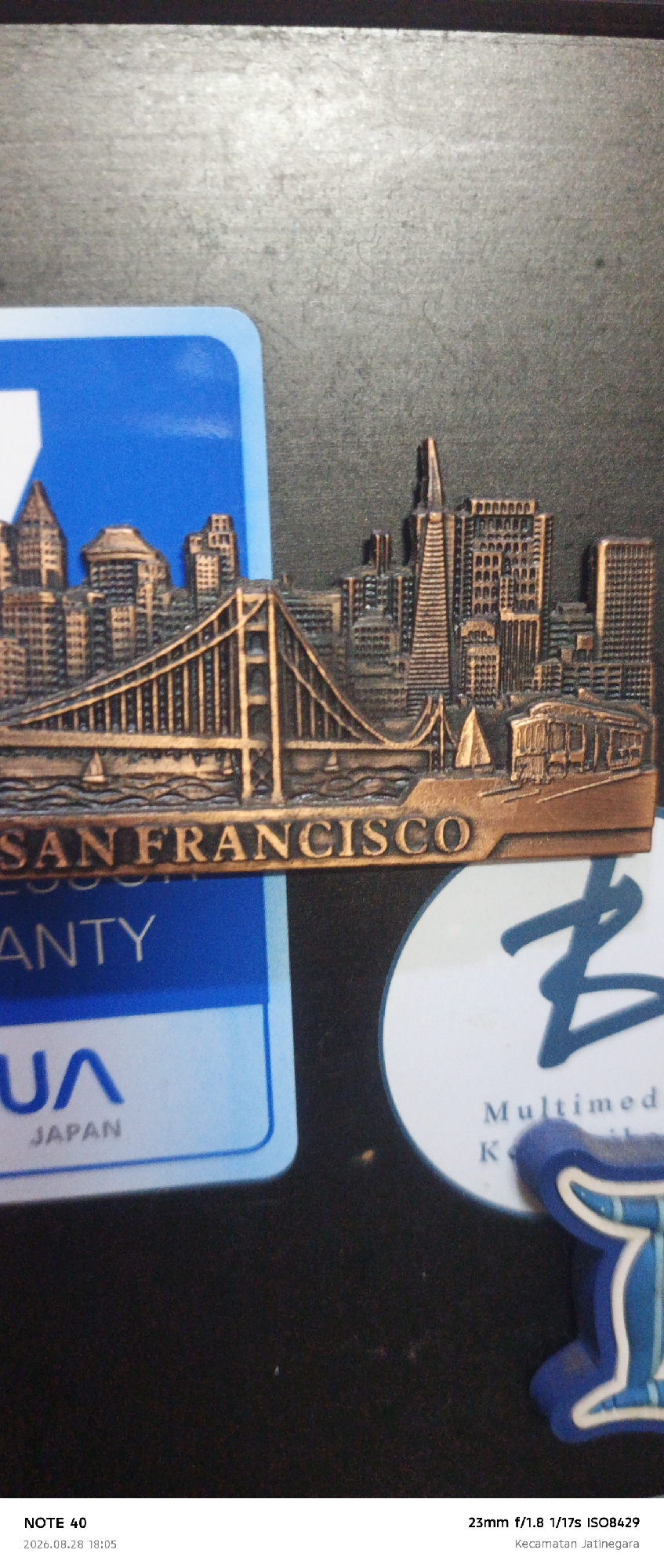 The image is a close-up, vertical shot of several objects on a dark, textured surface. In the foreground, on the left, is a portion of a blue and white rectangular sticker with the text "U/A JAPAN" visible. To the right of this sticker is a copper-colored miniature cityscape, featuring buildings and the Golden Gate Bridge, with the text "SAN FRANCISCO" below it. Further to the right, there is a white circular sticker with a dark blue stylized "B" logo and the text "Multimedia". Below this, partially obscured, is the letter "K" and another blue and white object that appears to be a foam craft item. The bottom of the image shows text overlays: "NOTE 40", "2026.08.28 18:05", "23mm f/1.8 1/17s ISO8429", and "Kecamatan Jatinegara". The presence of "Kecamatan Jatinegara" indicates the location is Jatinegara District in East Jakarta, Indonesia. The scene appears to be a flat lay of souvenirs or decorative items.