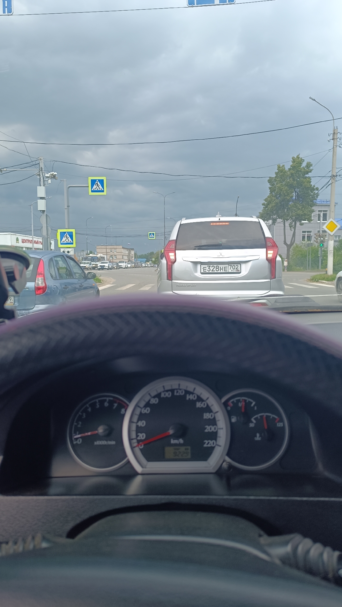 A first-person perspective shot from inside a vehicle shows a view through the windshield. The vehicle is stopped at an intersection with a traffic light visible in the distance. The dashboard instruments are visible in the foreground, with the speedometer indicating a speed of 0 km/h. Outside the vehicle, several cars are present, including a silver Mitsubishi Pajero Sport with a license plate reading E328HE 702. A pedestrian crossing sign is visible on the left side of the road. The sky is overcast with dark clouds, and trees and buildings line the street. The scene appears to be in Chaikovsky, Russia, suggested by the license plate's region code.