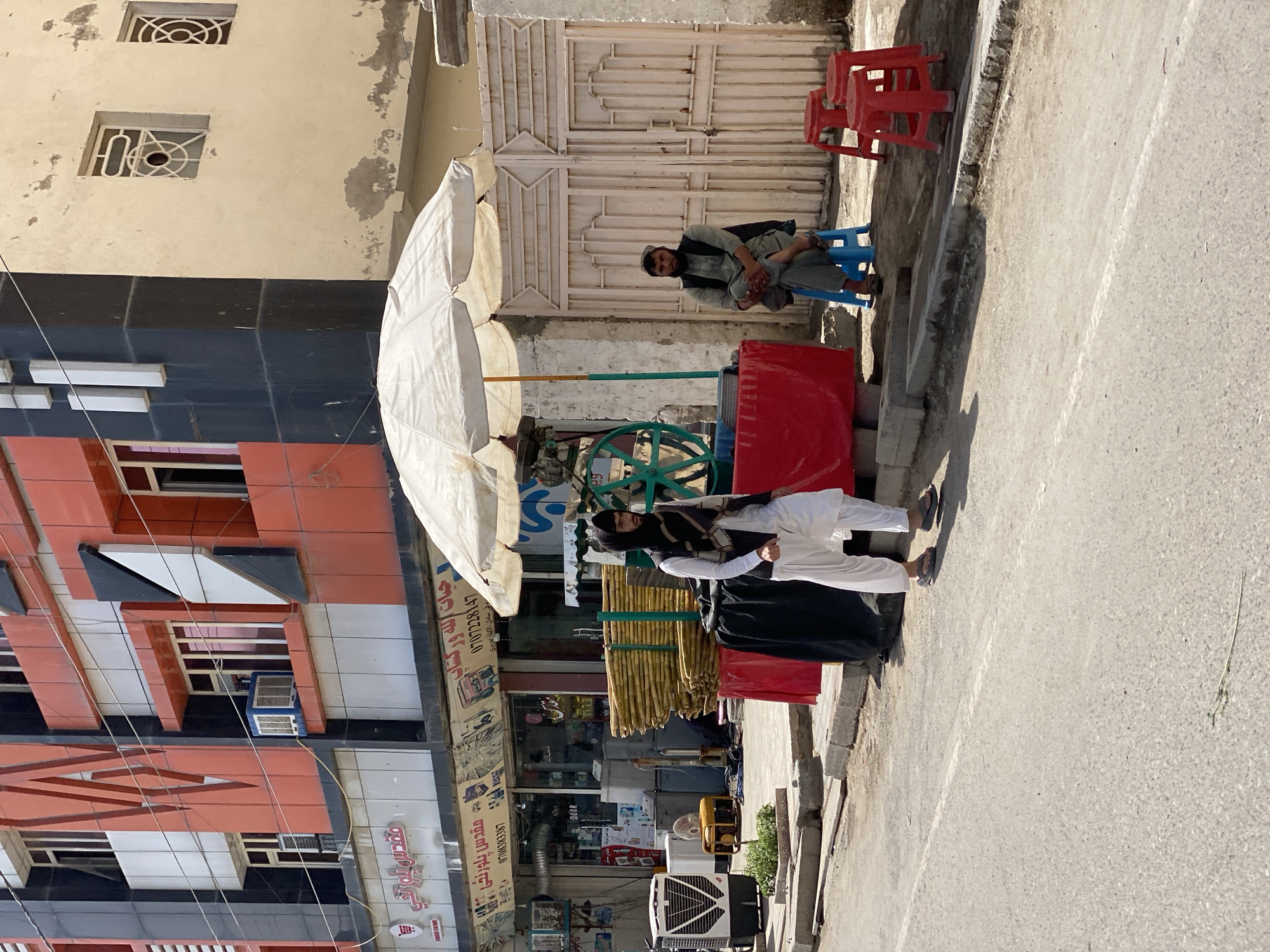 An outdoor scene on a street in Kandahar, Afghanistan features a sugar cane juice stand. Two men are present. One man, wearing a light gray tunic and vest with a cap, sits on a blue plastic stool in front of a closed beige gate. The second man, wearing a white shalwar kameez and a dark headscarf, stands near the juice stand, which has a red base and is covered by a large white umbrella. The stand has a green juicing machine. To the left of the stand are stacks of sugar cane stalks tied together. In the background, a multi-story building displays a modern facade with orange, black, and white geometric patterns. Shops are visible on the ground floor of the building. Air conditioning units are mounted on the building's exterior. The street is paved and appears to be in a commercial area.