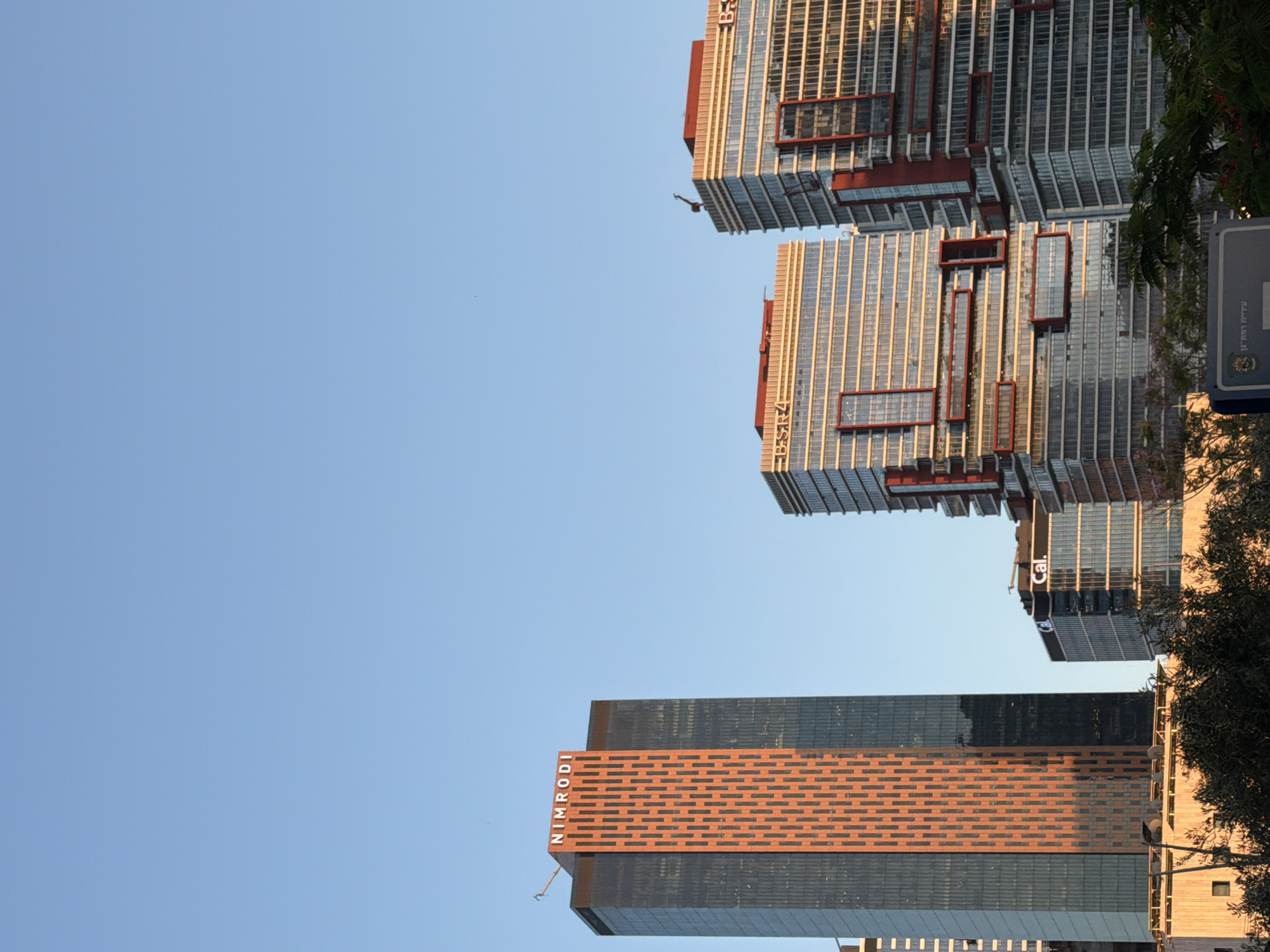 The image depicts the skyline of Bnei Brak, Israel, under a clear blue sky. The foreground is dominated by modern skyscrapers. The tallest building on the left features a distinctive orange patterned facade with the "NIMRODI" logo at its peak. To the right, two other prominent buildings with red accents and glass exteriors are visible, one of which has "B.S.R.4" branding. A smaller building with the "Cal." logo is positioned between the Nimrodi tower and the B.S.R. buildings. Trees and foliage are present in the lower part of the image, partially obscuring the base of some structures. The lighting suggests it is either early morning or late afternoon, casting a warm glow on the buildings.