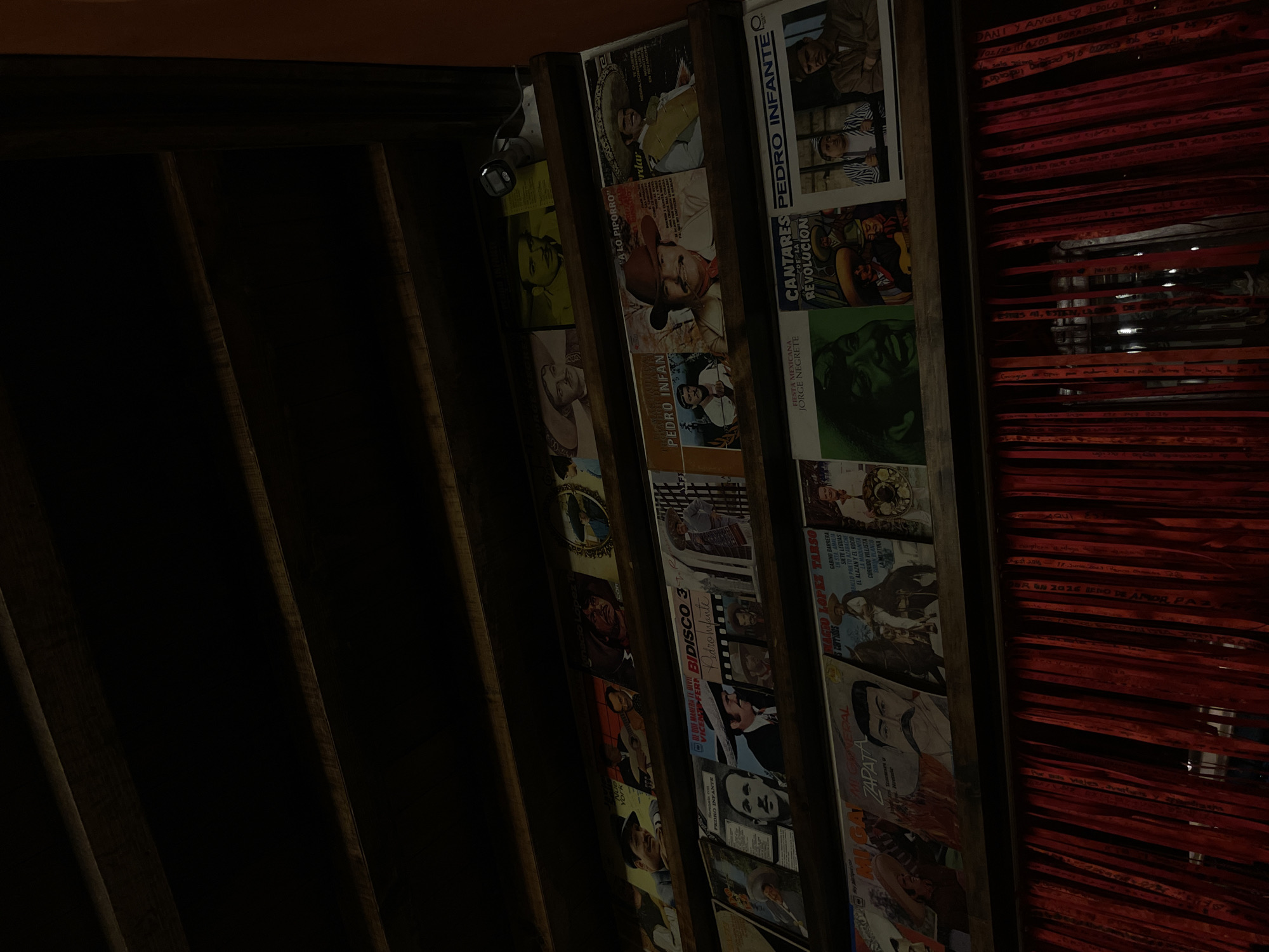 The image shows a close-up view of a dark interior, possibly a bar or cantina in Puebla, Mexico. The focus is on shelves filled with LP records, mostly of Mexican singers and actors like Pedro Infante, Jorge Negrete, and covers of the "Corridos." Adjacent to the records are numerous red ribbons, each bearing visible names or inscriptions. The dim lighting and tight framing suggest an intimate, possibly nostalgic setting. In the upper left, there is a partial view of dark wooden beams.