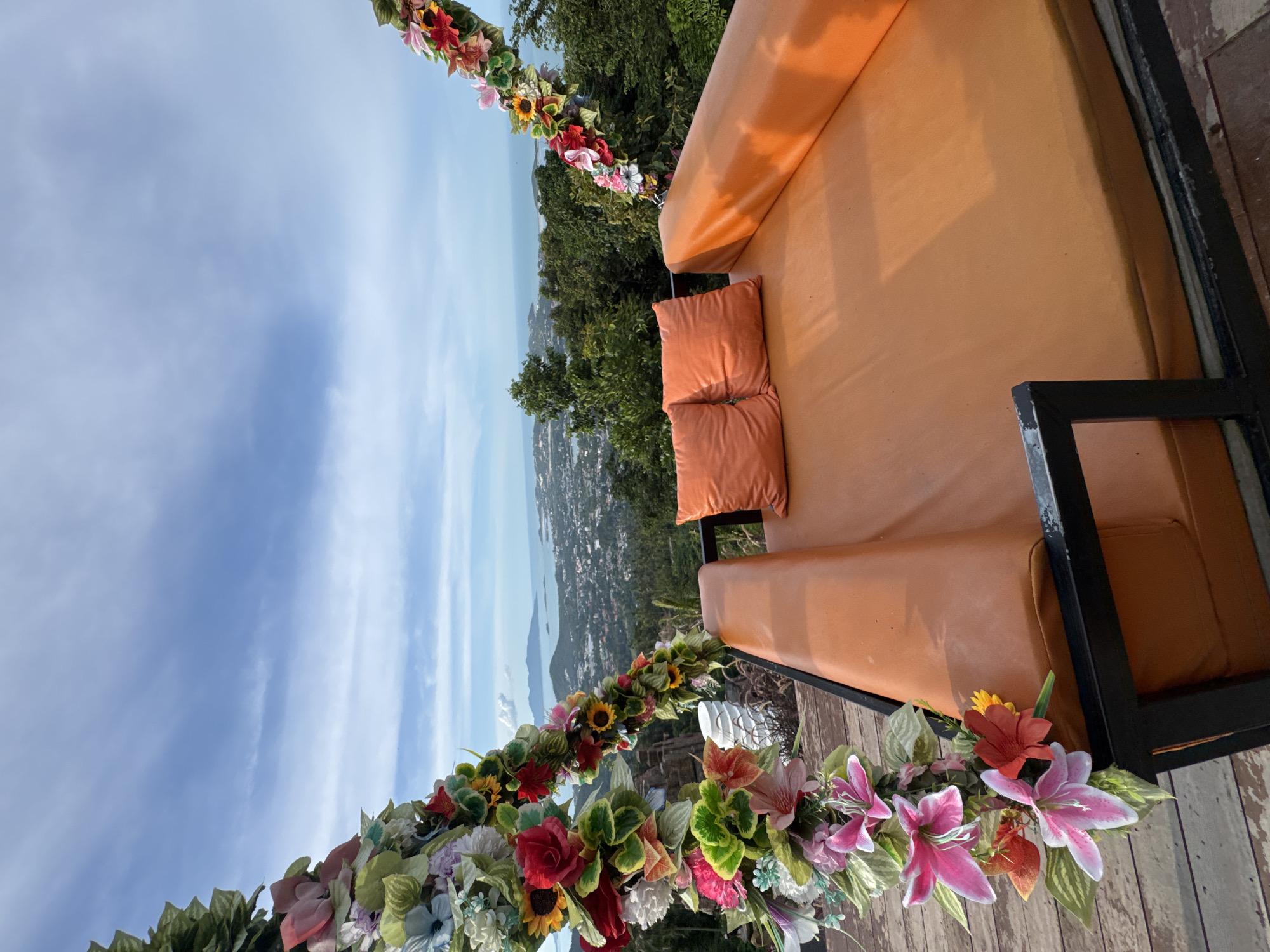 An outdoor daybed with orange cushions and two orange pillows is positioned on a wooden deck, overlooking a city and ocean landscape. The daybed is framed by two decorative arches adorned with artificial flowers and greenery. The sky is blue with scattered clouds. The city appears to be coastal, with buildings nestled among lush green trees leading down to the water. The overall scene suggests a scenic viewpoint or a resort setting.