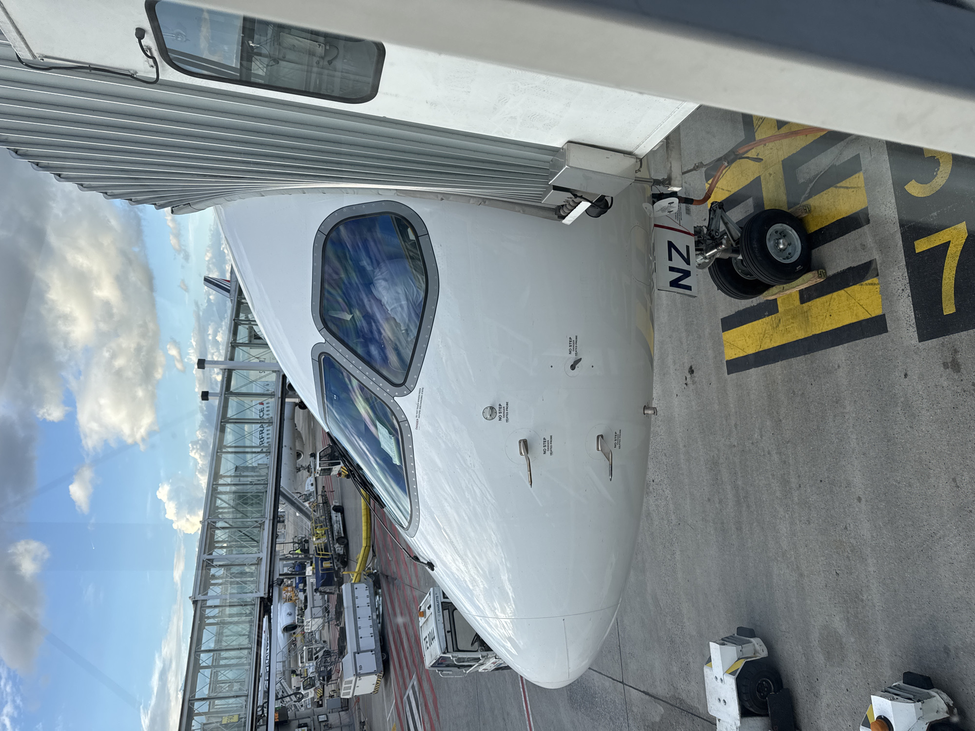 The image shows the nose of an Air New Zealand (NZ) aircraft parked at a gate, likely in Le Mesnil-Amelot, France, given the Air France gate visible in the background. The aircraft is connected to a jet bridge. Ground equipment, including tugs, are visible near the nose gear. The markings on the ground indicate the aircraft's position at gate 7. The sky with clouds is visible through the jet bridge windows.