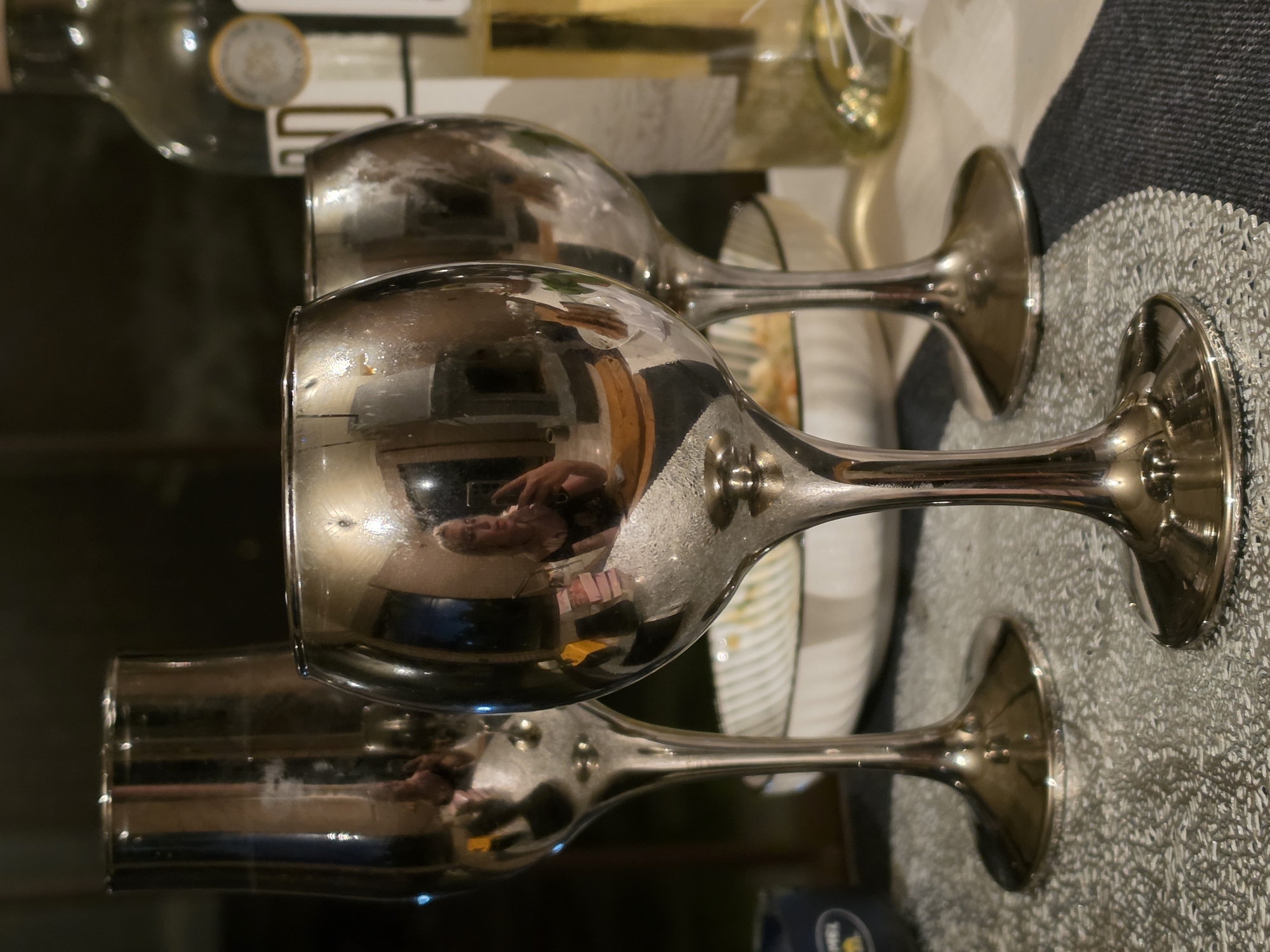 The image displays three metallic wine glasses arranged on a table. The glasses are highly reflective, capturing distorted images of the surrounding environment. In the central glass, a reflection shows a person taking a photograph with a smartphone, holding it up to capture their own image. Behind the glasses, a bottle of wine is visible. The overall setting appears to be an indoor environment, possibly a home or a restaurant, with a dark background. There is no specific information in the image to indicate the location is in Nagorny, Russia.