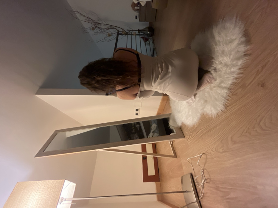 The image depicts the interior of a residence, located in Cava de' Tirreni, Italy. A single female individual is observed from behind, kneeling on a white faux fur rug. Her hair is dark brown and curly, extending to her shoulders. She wears a light-colored, off-the-shoulder dress, with a dark bra visible beneath, and her feet are bare. To her left, a freestanding, full-length mirror with a light wooden frame is positioned. The mirror's reflection shows an interior space, including what appears to be a bed with white linens. A silver-toned floor lamp with a rectangular fabric shade stands on the far left, providing illumination; its power cord is visible on the light wooden floor. The walls are white. Two electrical outlets are present on the wall between the mirror and the individual. In the background, to the right, a dark metal railing and a dark vase containing decorative branches are visible, alongside a partially obscured light-colored dresser or table. The room features light wooden flooring throughout, and the overall lighting is dim and warm.
