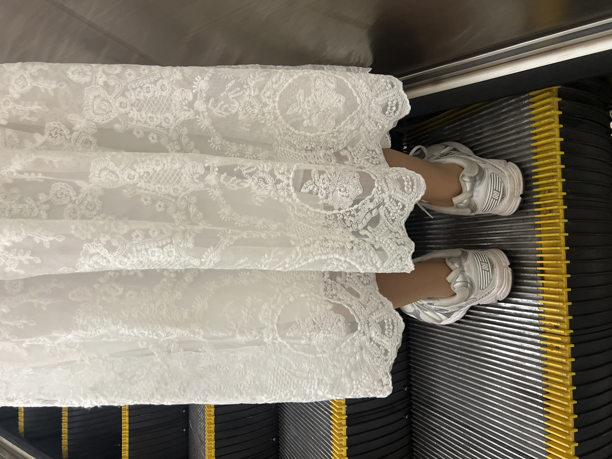 A downward-angled shot focuses on a person's feet and the lower portion of their attire, set against the backdrop of an escalator. The individual is wearing a long, white lace dress that extends over the majority of the frame. Their feet are shod in white and silver athletic shoes, positioned on the black and yellow steps of the escalator. The escalator is moving; its direction is unclear. The lighting is bright and appears to be artificial, indicating an indoor setting, though it is difficult to pinpoint an exact location in Saint Petersburg, Russia.